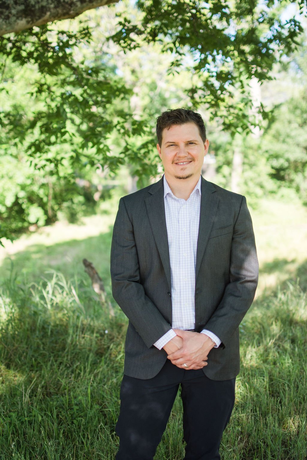 A man in a dark gray suit and light-colored shirt standing outdoors in a park with green trees and grass, smiling.