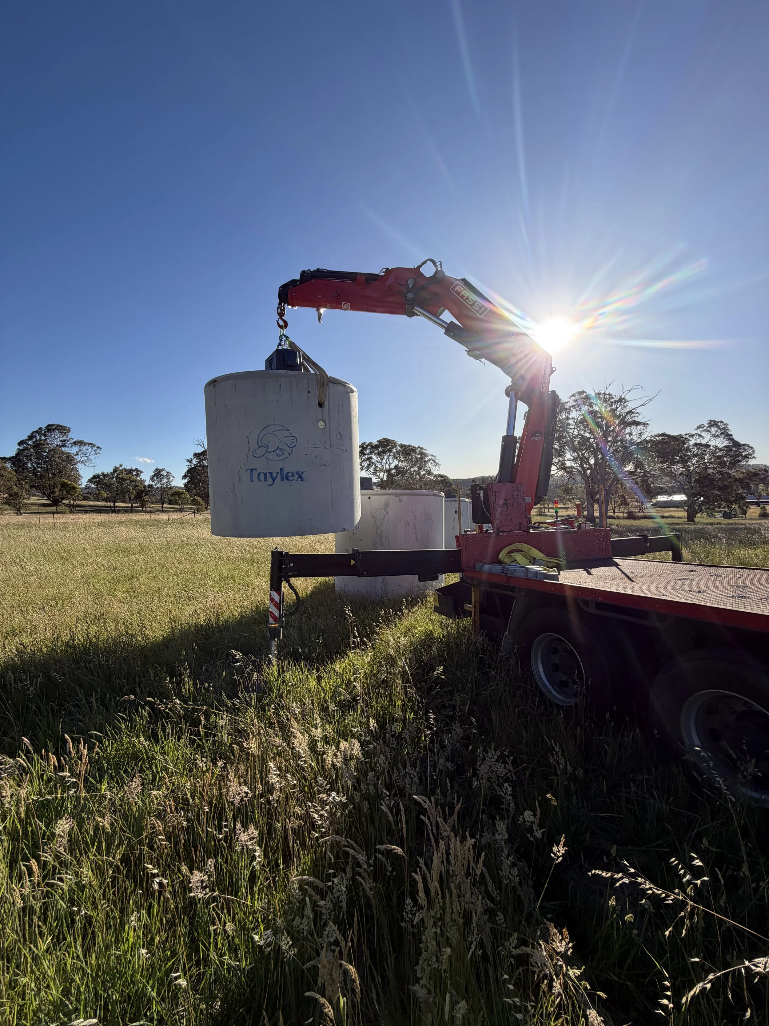 A red crane on a trailer lifting a white cylindrical tank with a logo and text, set in a grassy field with trees and clear blue sky, sunlight creating lens flare.