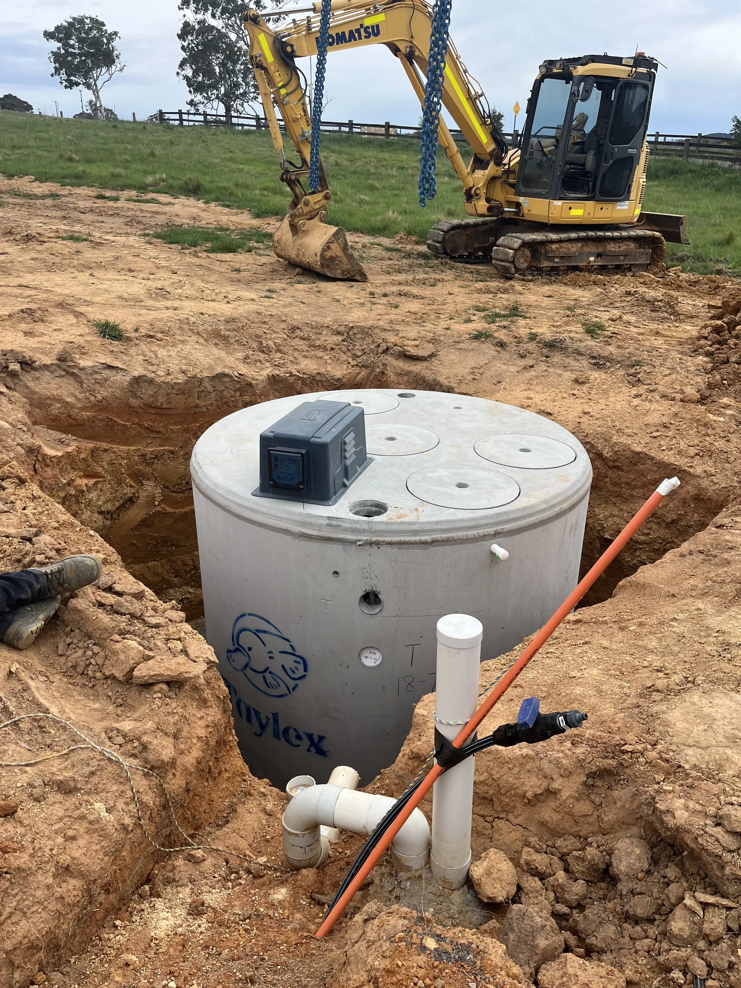 Underground utility installation site showing a large white cylindrical tank labeled Lwyflex, partially buried in a trench with PVC piping, connecting to various pipes and fittings. In the background, a small yellow excavator is parked on a grassy hill.