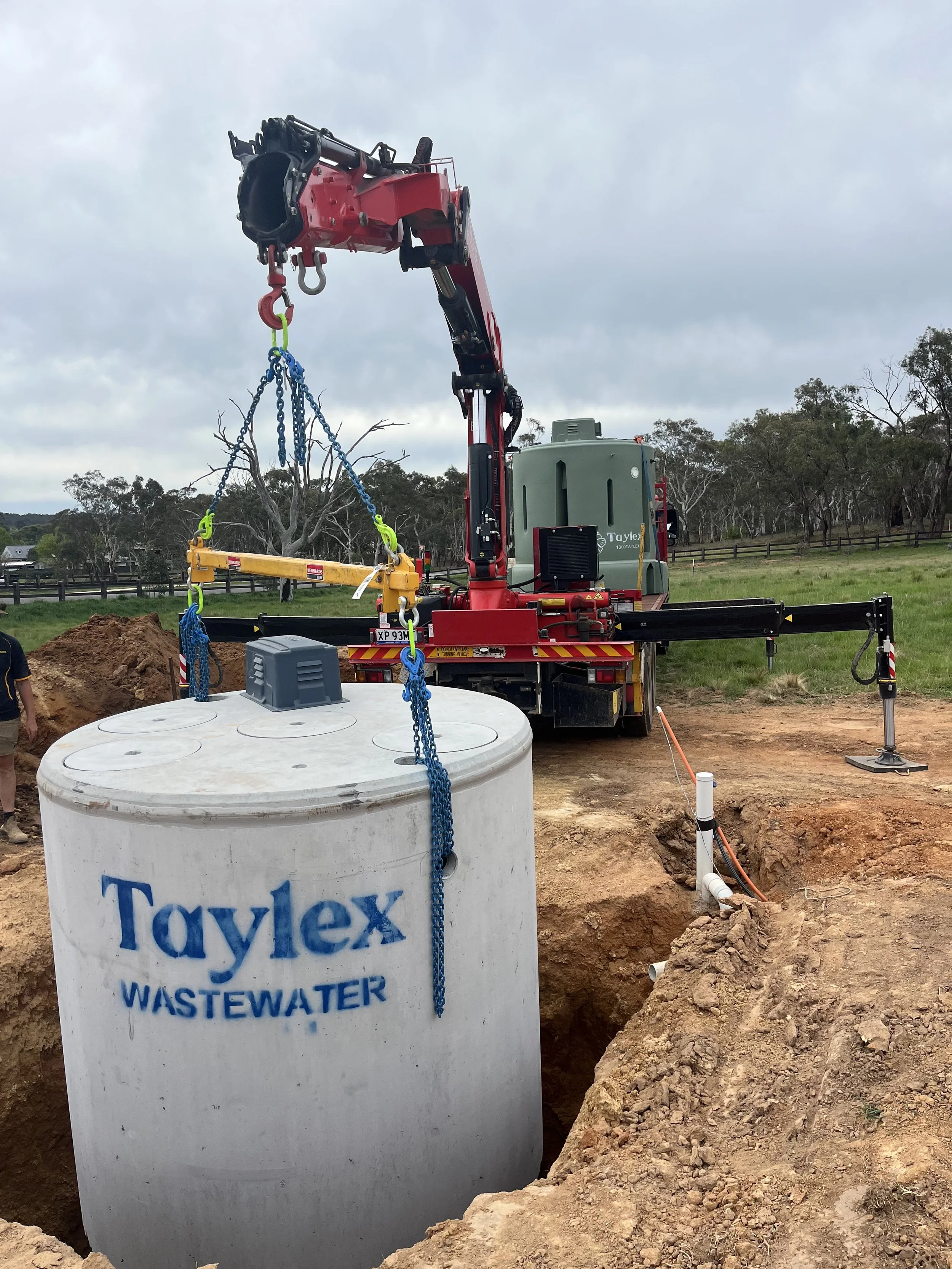 A construction site where a red crane is installing a large underground waste water tank labeled 'Townley Wastewater.' The tank is partially buried in the ground, with exposed dirt and pipes nearby.
