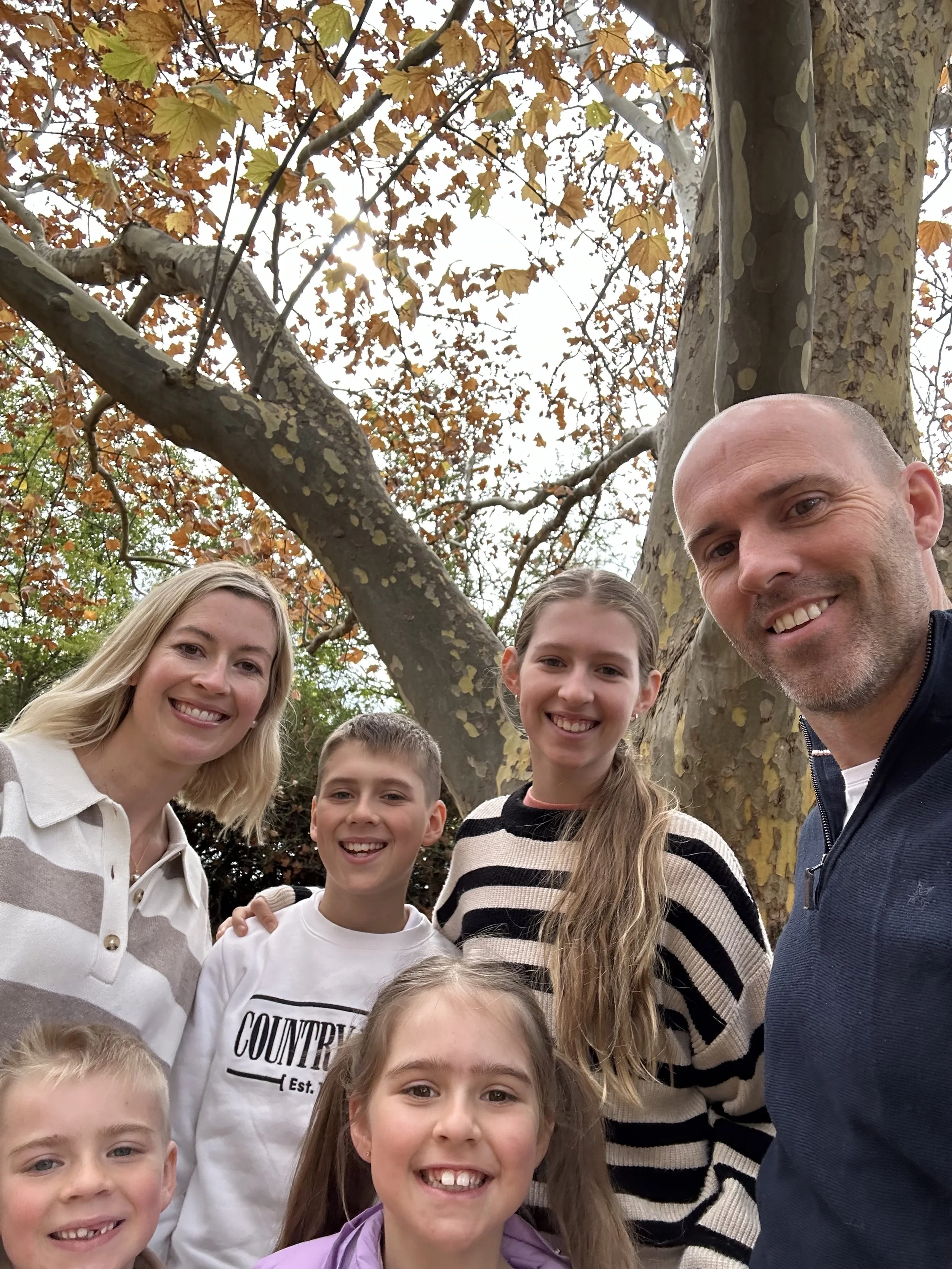A group of people, including children and adults, smiling and taking a selfie outdoors in front of a large tree with autumn-colored leaves.
