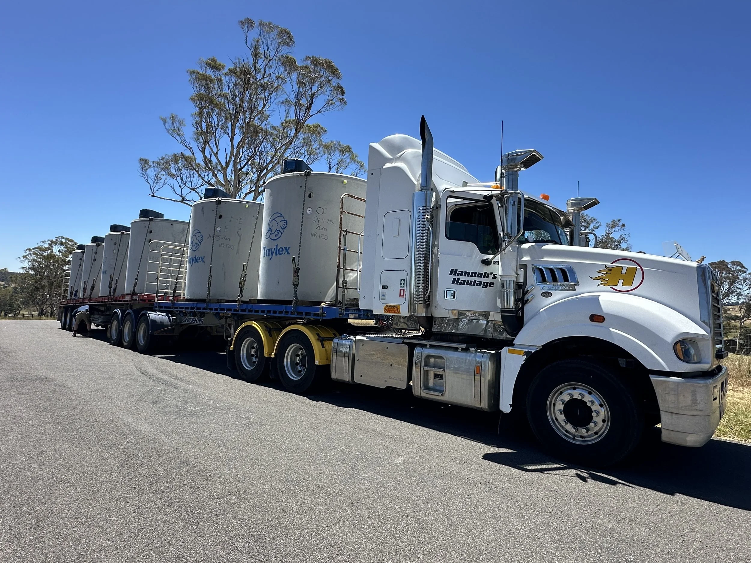 A large white semi-truck with a flat trailer carrying several white tanks labeled Taylex, parked on a paved road under a clear blue sky. Trees are visible in the background.