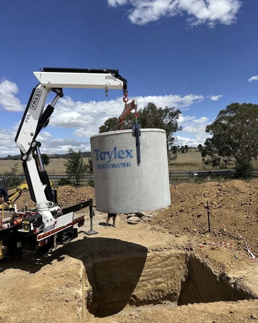 A crane lifting a large concrete wastewater tank labeled 'Taylex WASTEWATER' during installation at a construction site with a dirt and gravel ground, trees, and a blue sky with scattered clouds in the background.