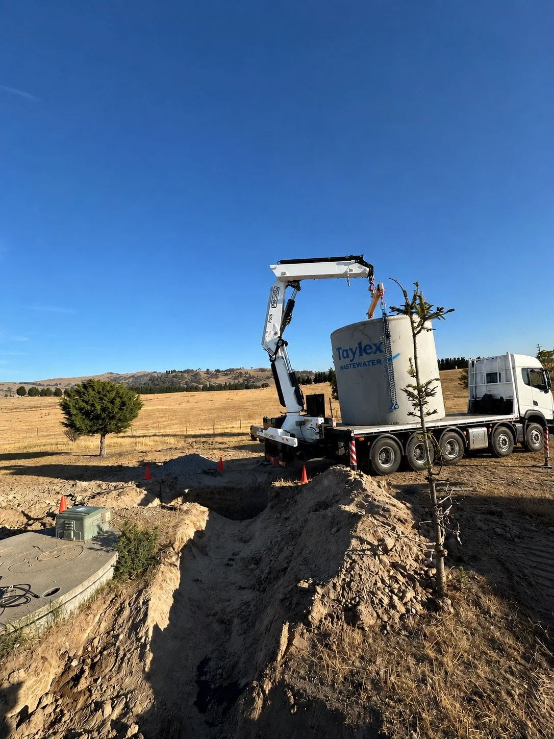 A construction site on a cloudy day showing a truck with a crane lifting a large wastewater tank labeled 'Taylex WASTEWATER' into place. There are small trees, orange cones, and a trench in the dirt.