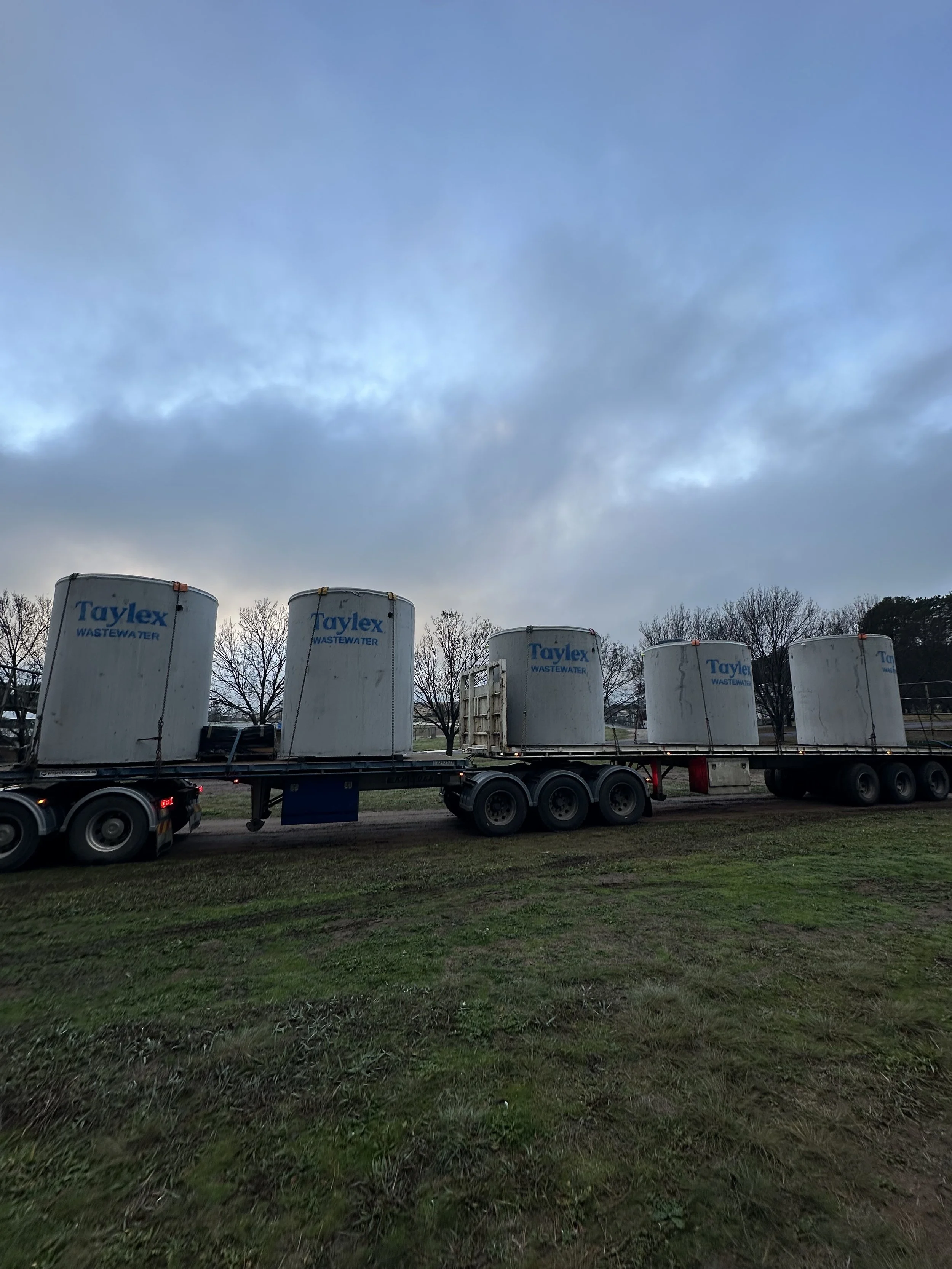 Large white industrial tanks labeled 'Taylex Wastewater' on a flatbed truck, parked on grass with leafless trees and a cloudy sky in the background.