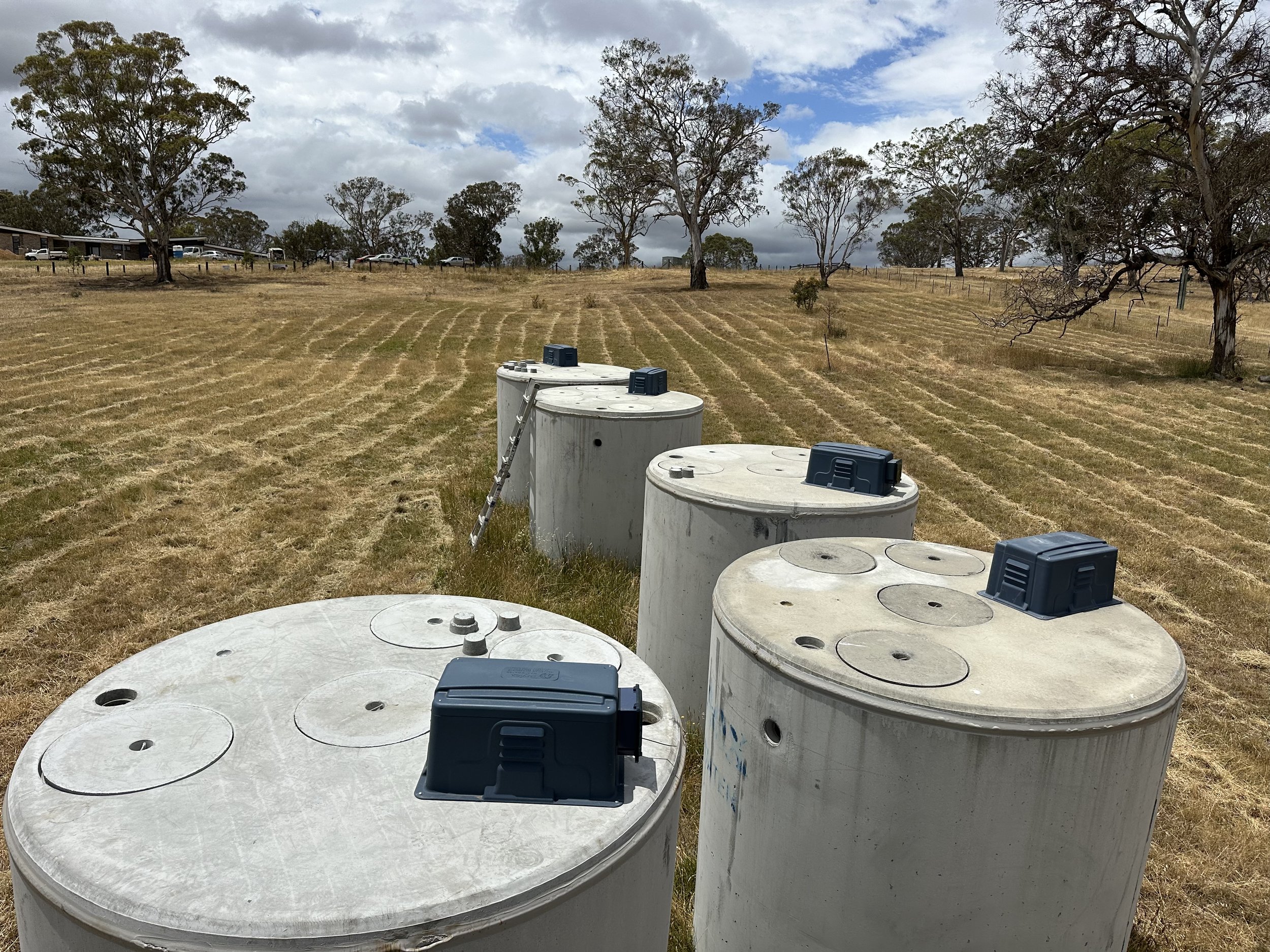 Open water storage tanks in a grassy field with scattered trees, cloudy sky.
