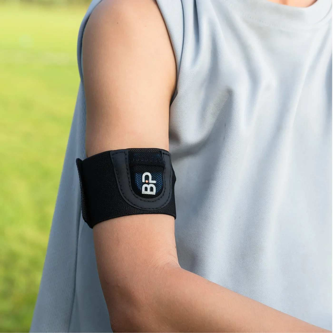 A person's upper arm with a black fitness tracker or armband with the letters 'BP' on it, against a blurred outdoor background.