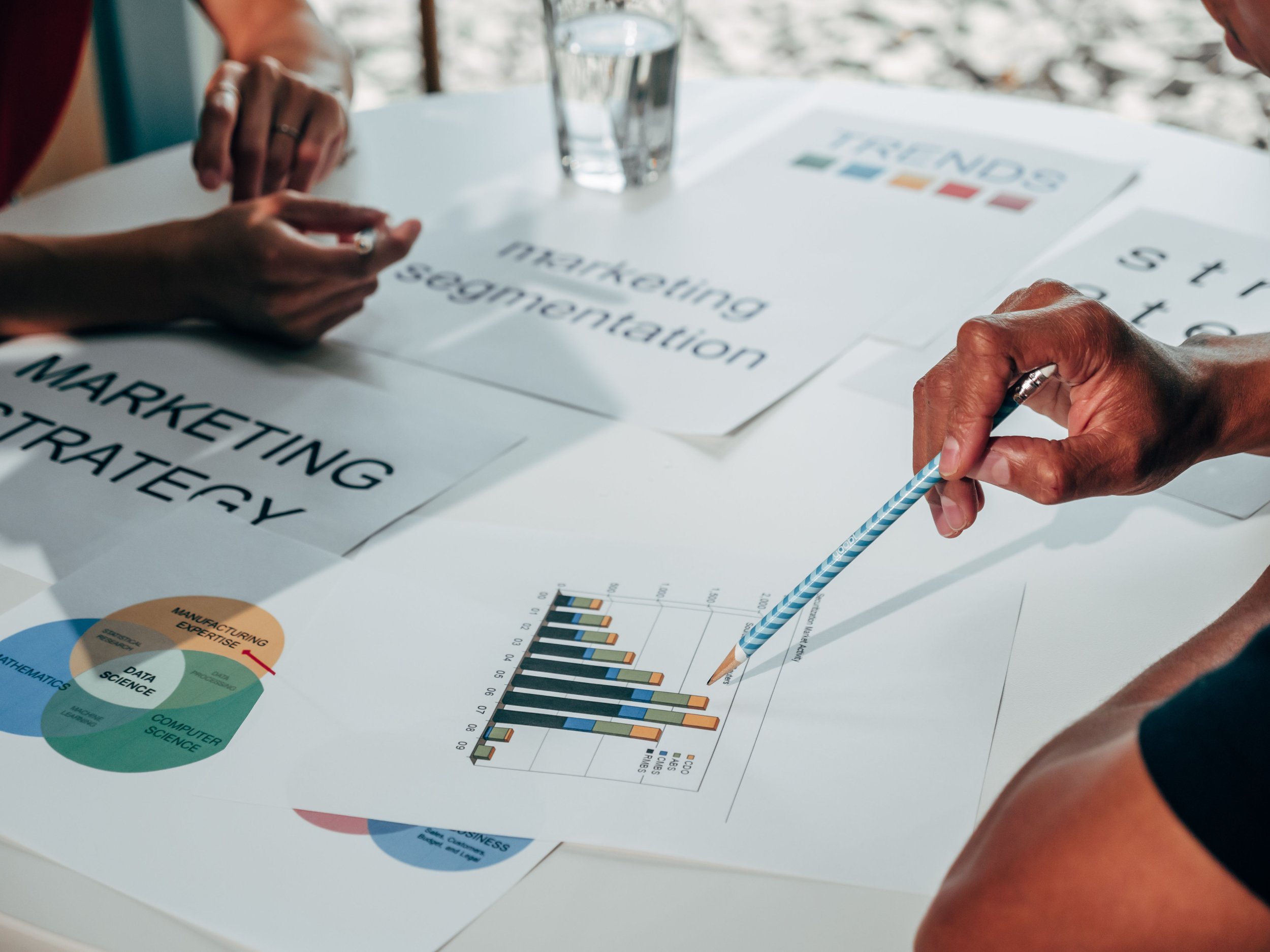 Close-up of two people working on printed business or marketing materials featuring graphs and charts, with a glass of water on the table.