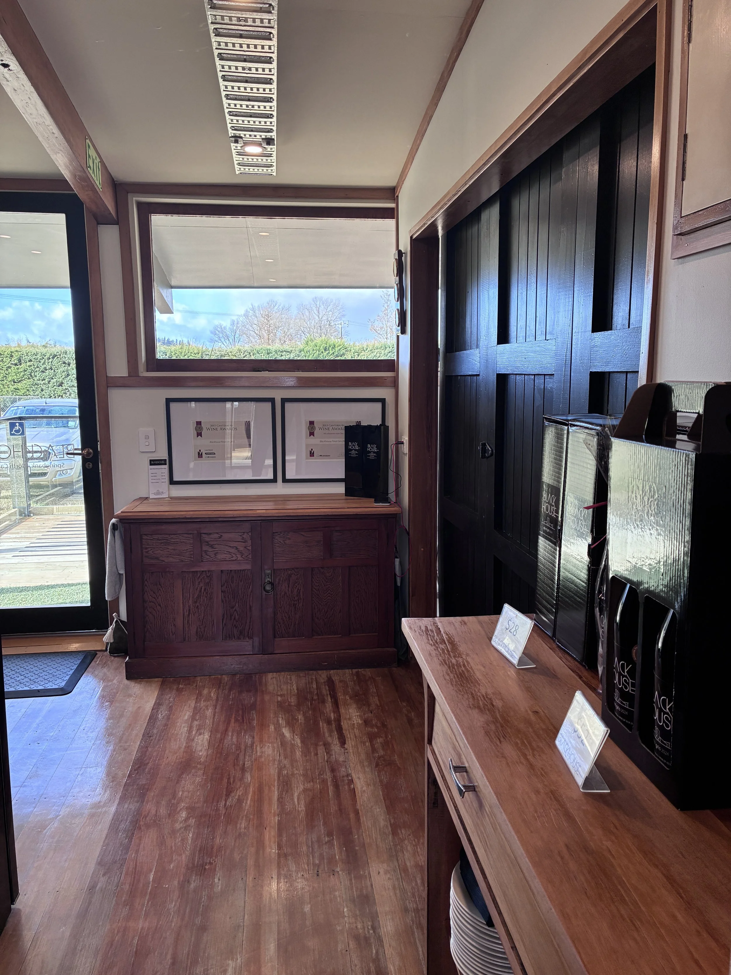 Interior of the cellar door with wooden furniture, including a small table and cabinets, and displays with wine bottles on a counter, with a large window showing trees outside.
