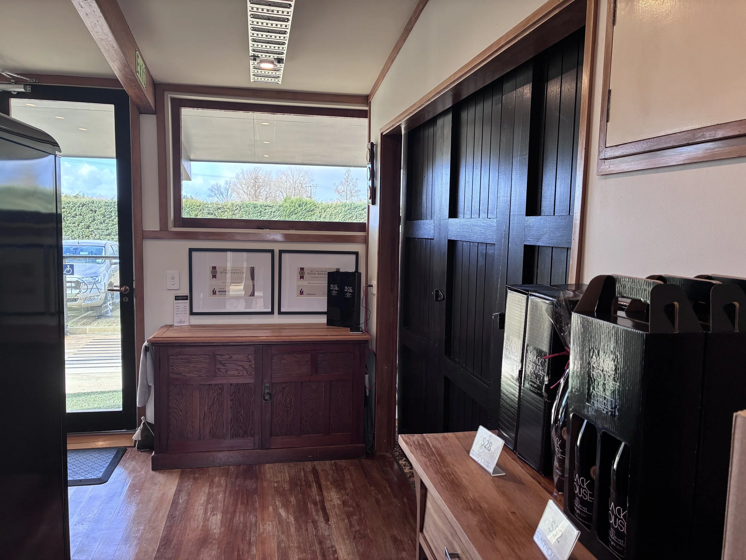 Interior of a store with wooden floors and walls, featuring a wooden counter, framed certificates or awards on the wall, and shelves displaying black boxes of Blackhouse wine.