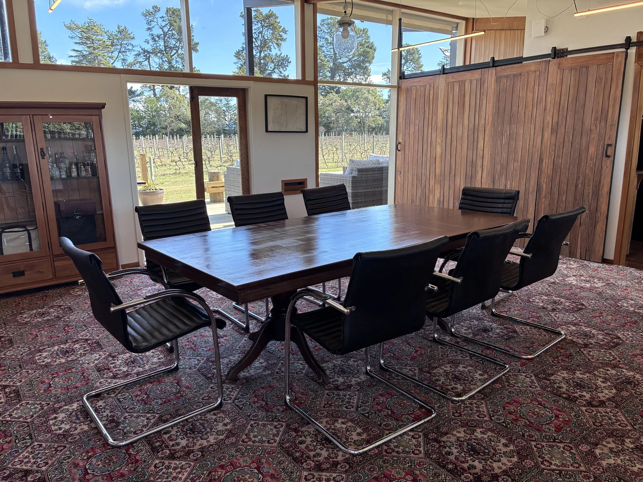 A conference room with a large wooden table surrounded by black chairs, a glass cabinet on the left, wooden barn doors hide the kitchenette, and large windows showing a vineyard and trees outside.
