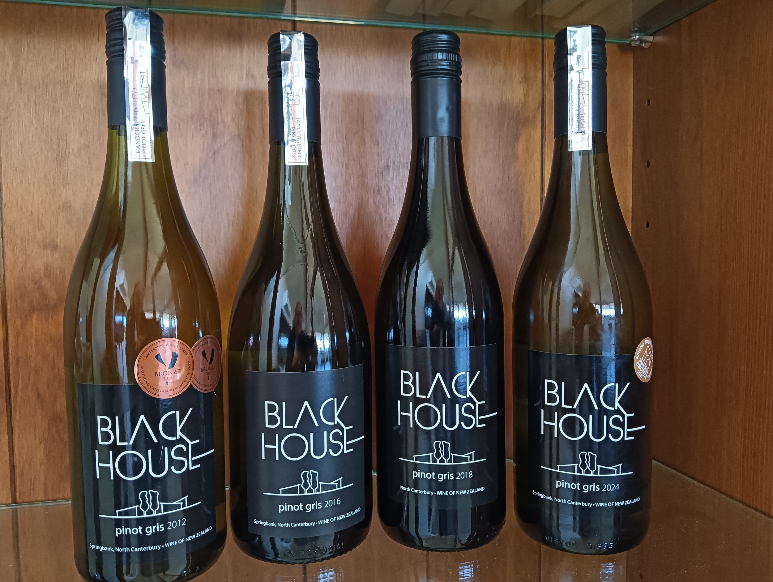 Four wine bottles from Black House winery arranged in a row inside a wooden display case. The bottles have black labels with white text indicating different years: 2012, 2016, 2018, and 2024, featuring Pinot Gris from North Canterbury, New Zealand.