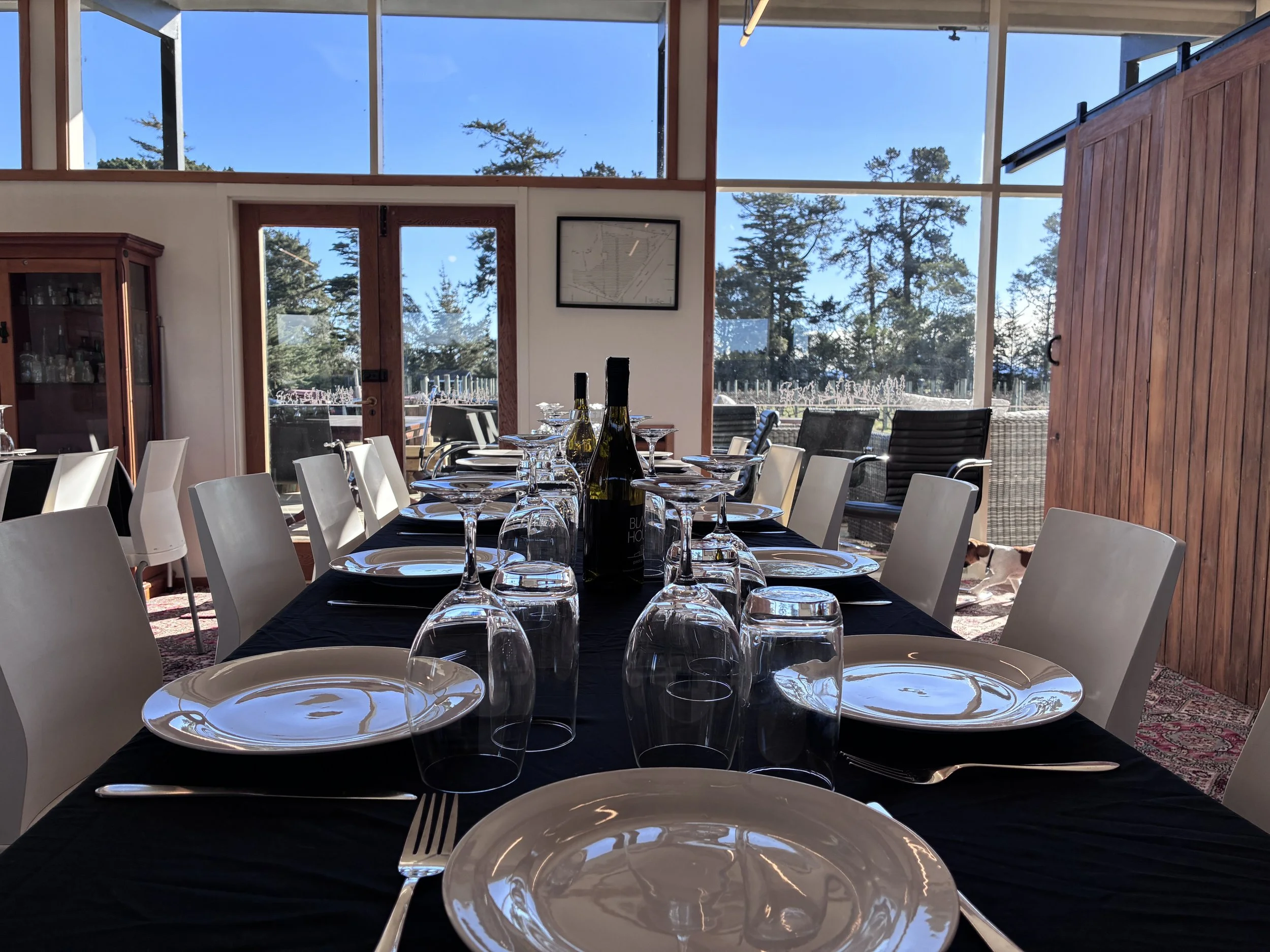 A dining area with a long table set with white plates, wine glasses, and bottles of wine. Large windows reveal trees and blue sky outside. The room has wooden accents and white chairs.
