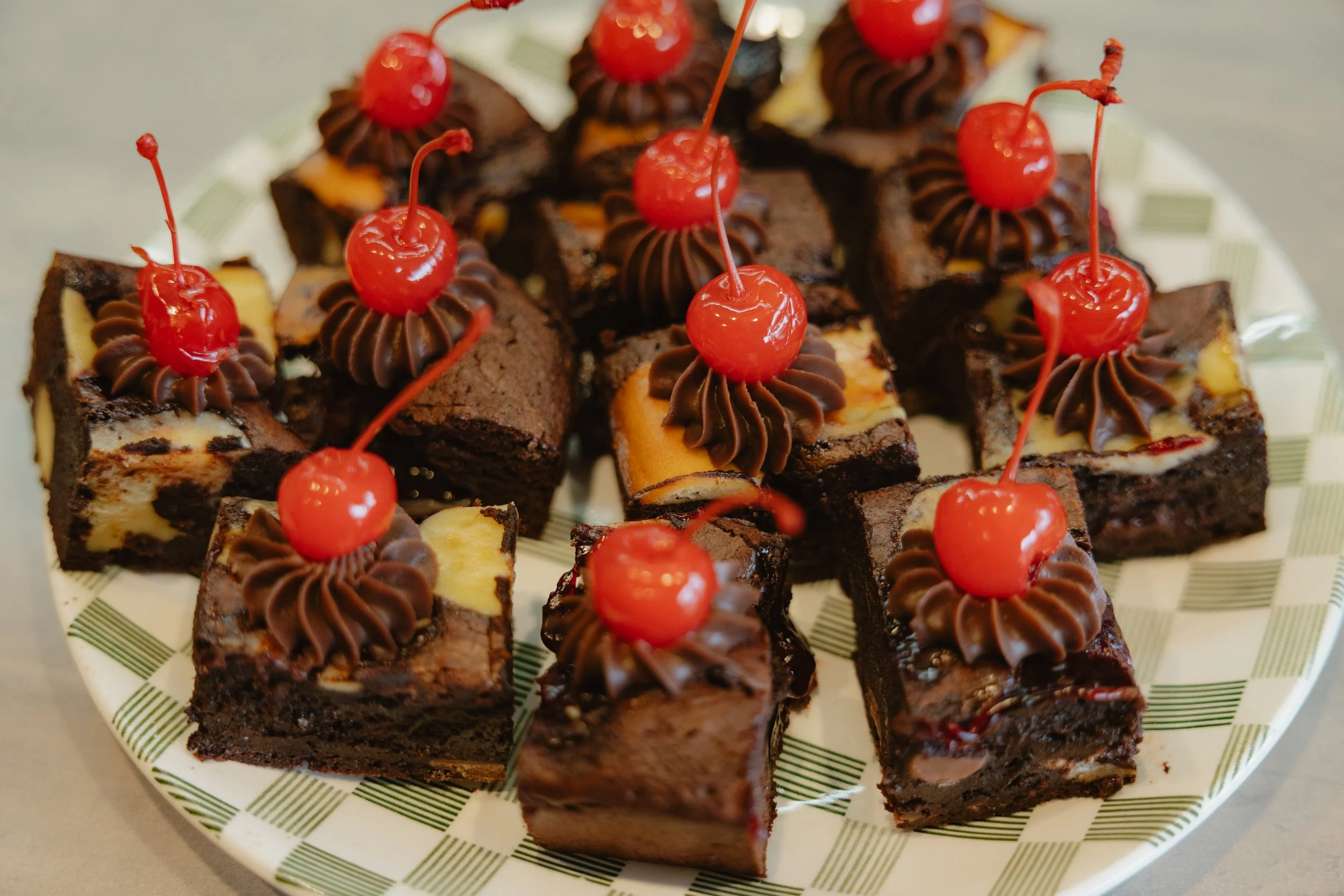 Close-up of small chocolate brownie squares topped with chocolate frosting and maraschino cherries on a white plate with a green striped pattern.