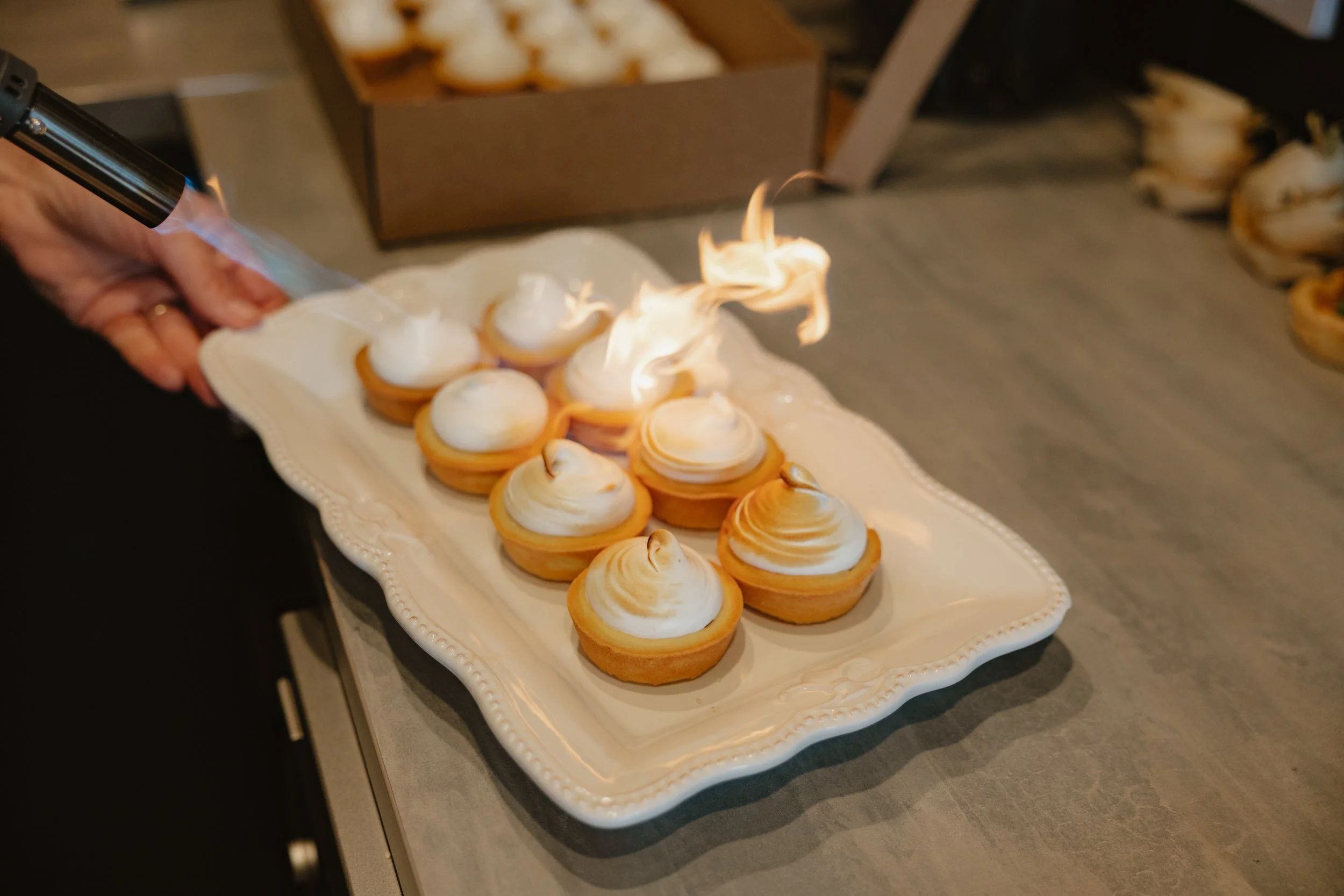 A white decorative platter with small open tarts topped with toasted meringue, one tart on the left is on fire with a small flame, held by a person's hand.