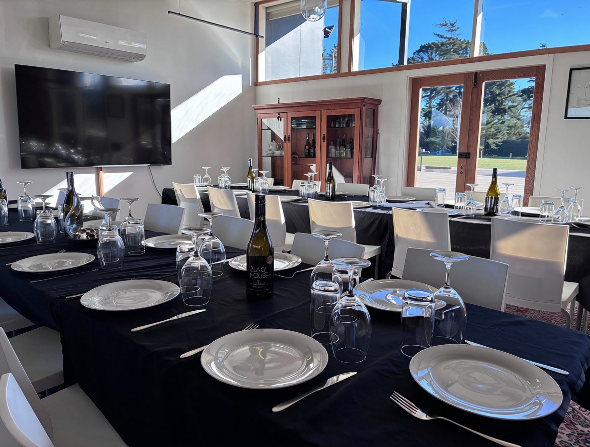 Indoor dining setup with black tablecloths, white plates, wine bottles, glasses, and silverware, in a bright room with large windows and a view of trees outside.