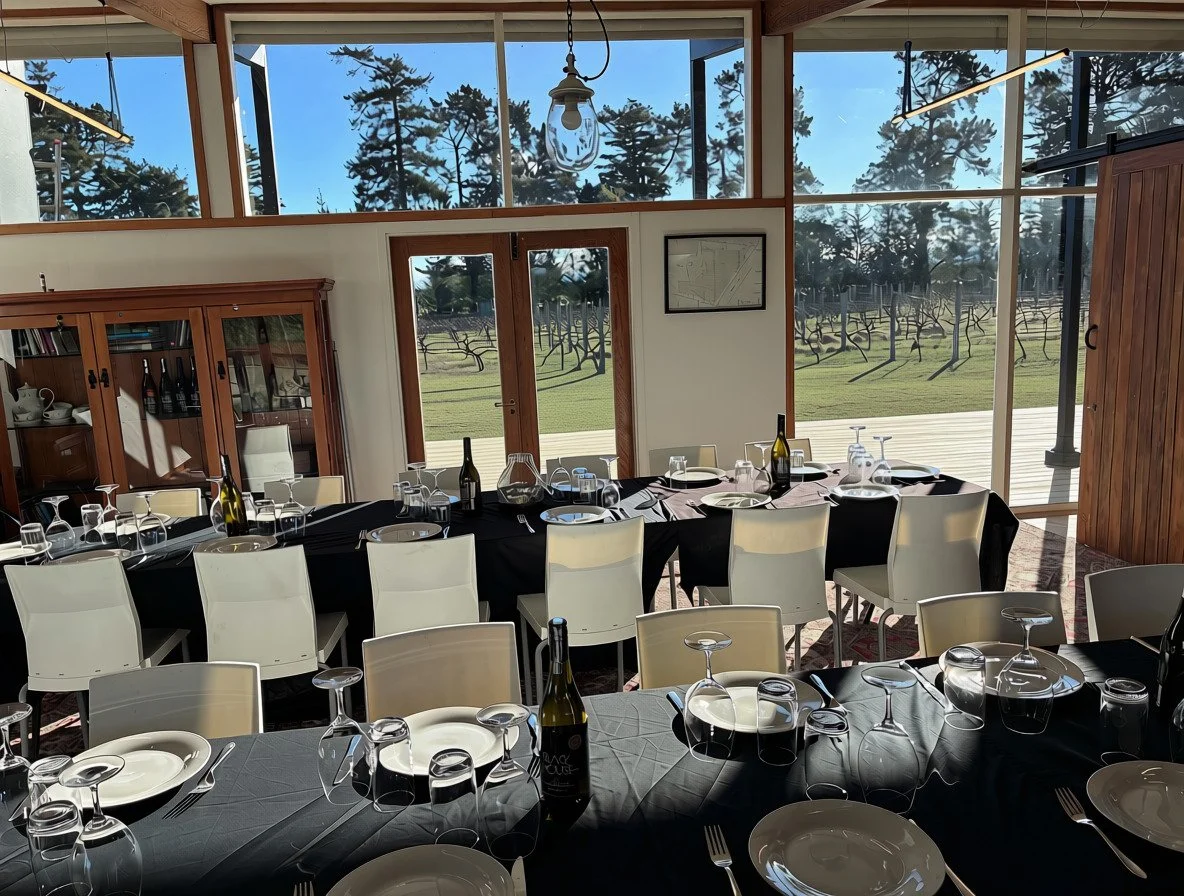 A dining room with large windows showing trees outside. The room has two tables set with black tablecloths, plates, wine bottles, and glasses, ready for a meal.