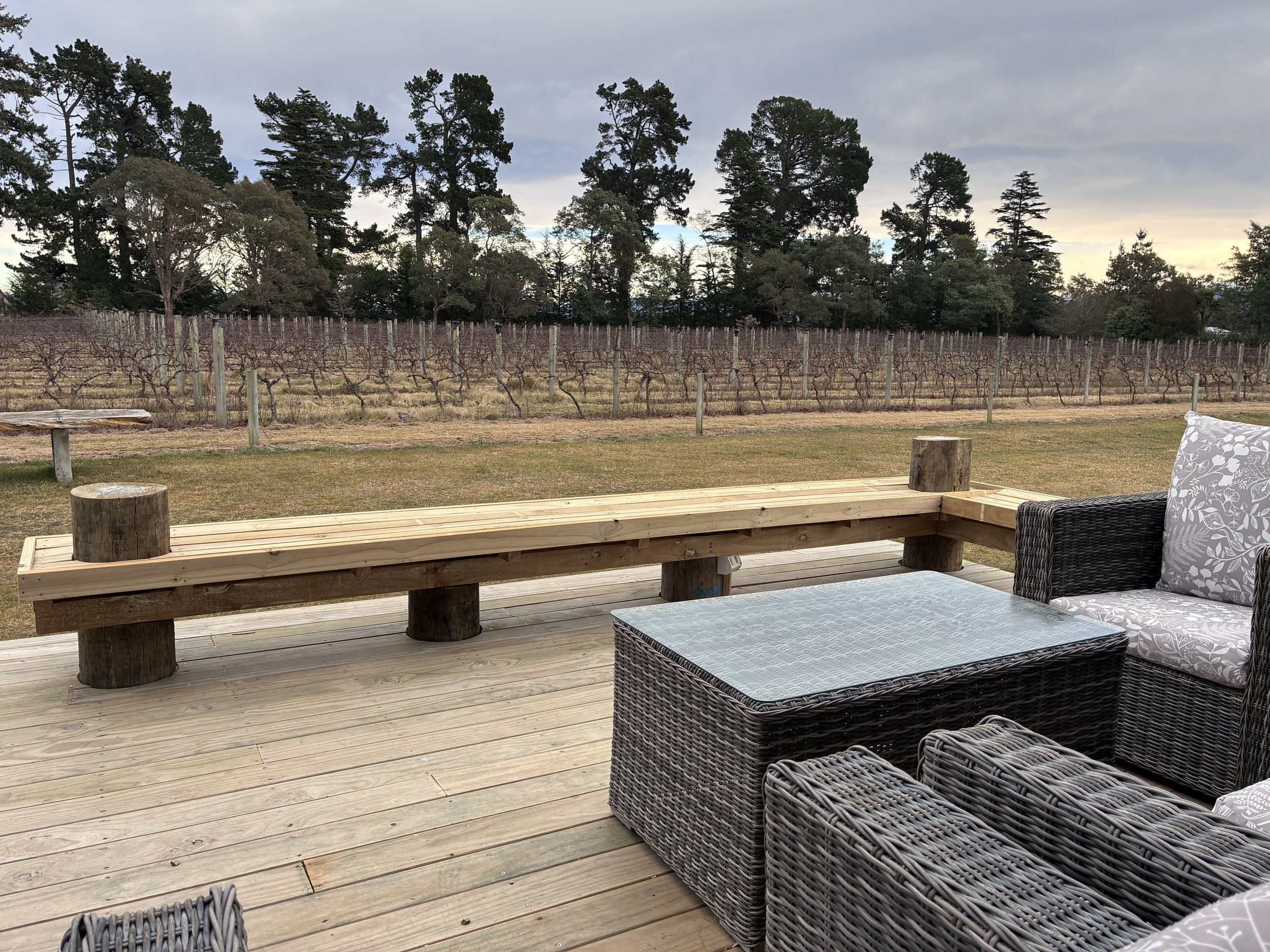 Deck with wicker furniture, a wooden deck, agricultural vineyard with leafless vines, tall trees in the background, and cloudy sky.