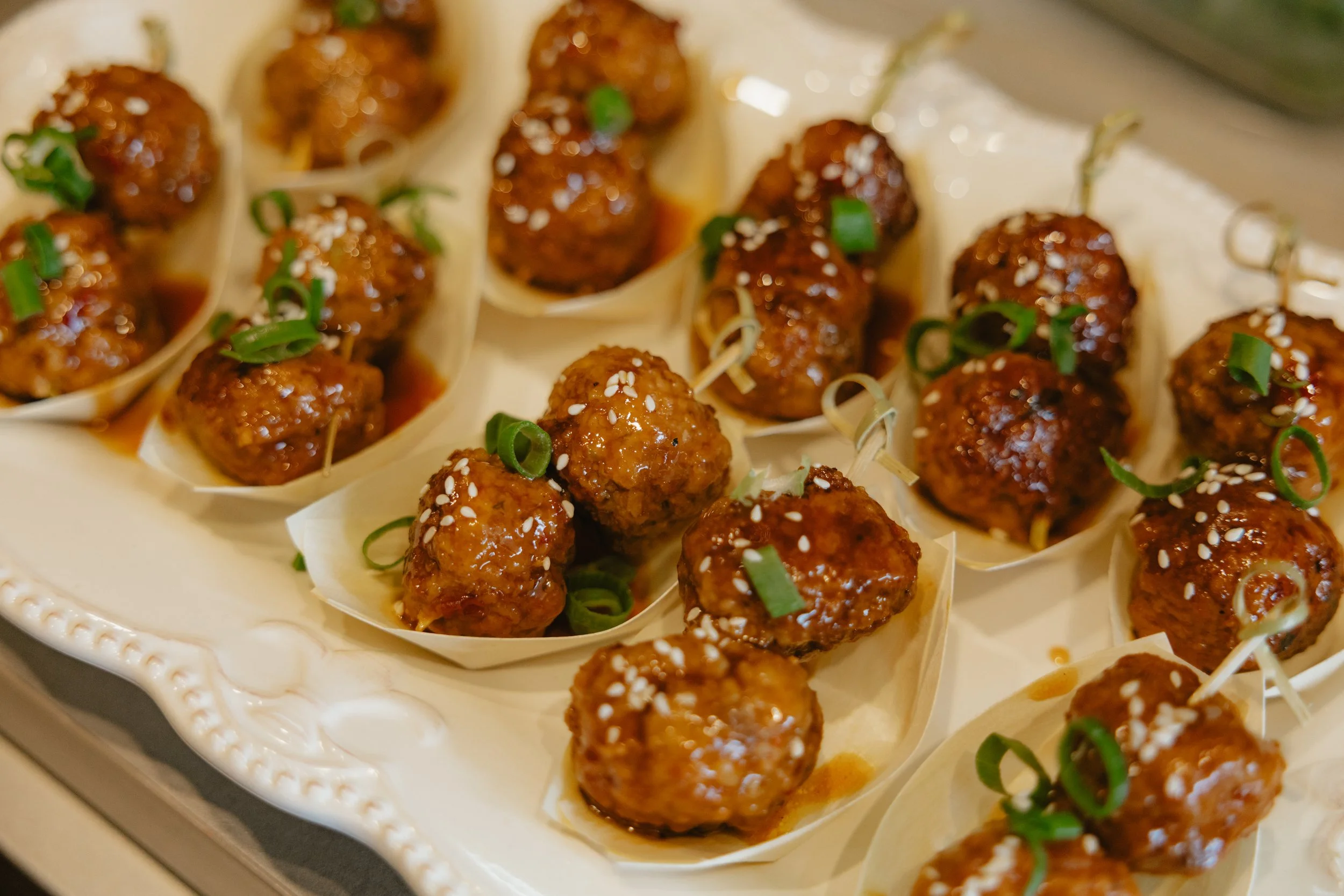 Close-up of several meatballs topped with chopped green onions and sesame seeds, served in paper cups on a decorative platter.