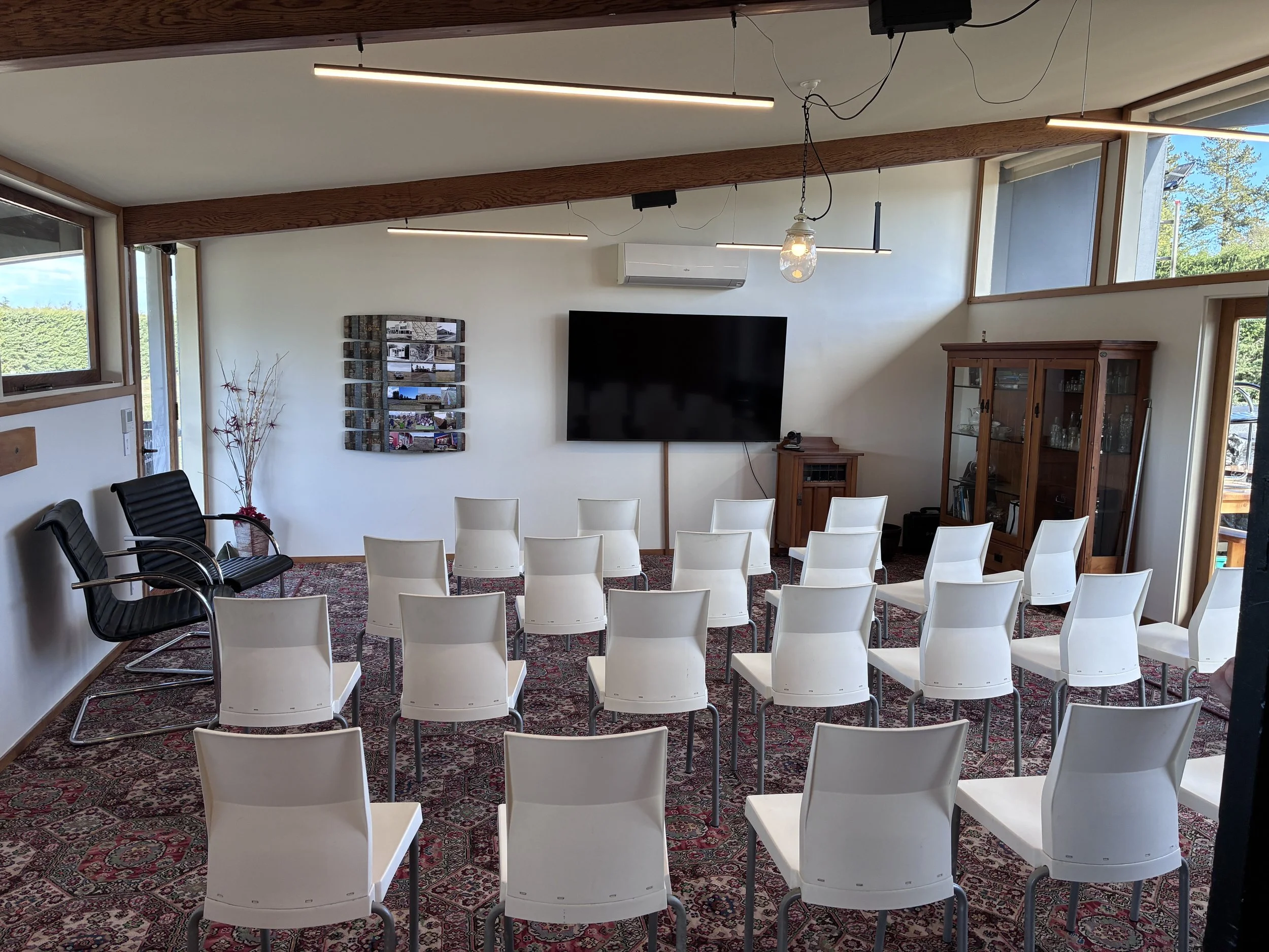 Interior of a room set up for a presentation or meeting with white chairs arranged in rows, a wall-mounted TV, artwork on the wall, a cabinet with glass doors, and large windows letting in natural light.