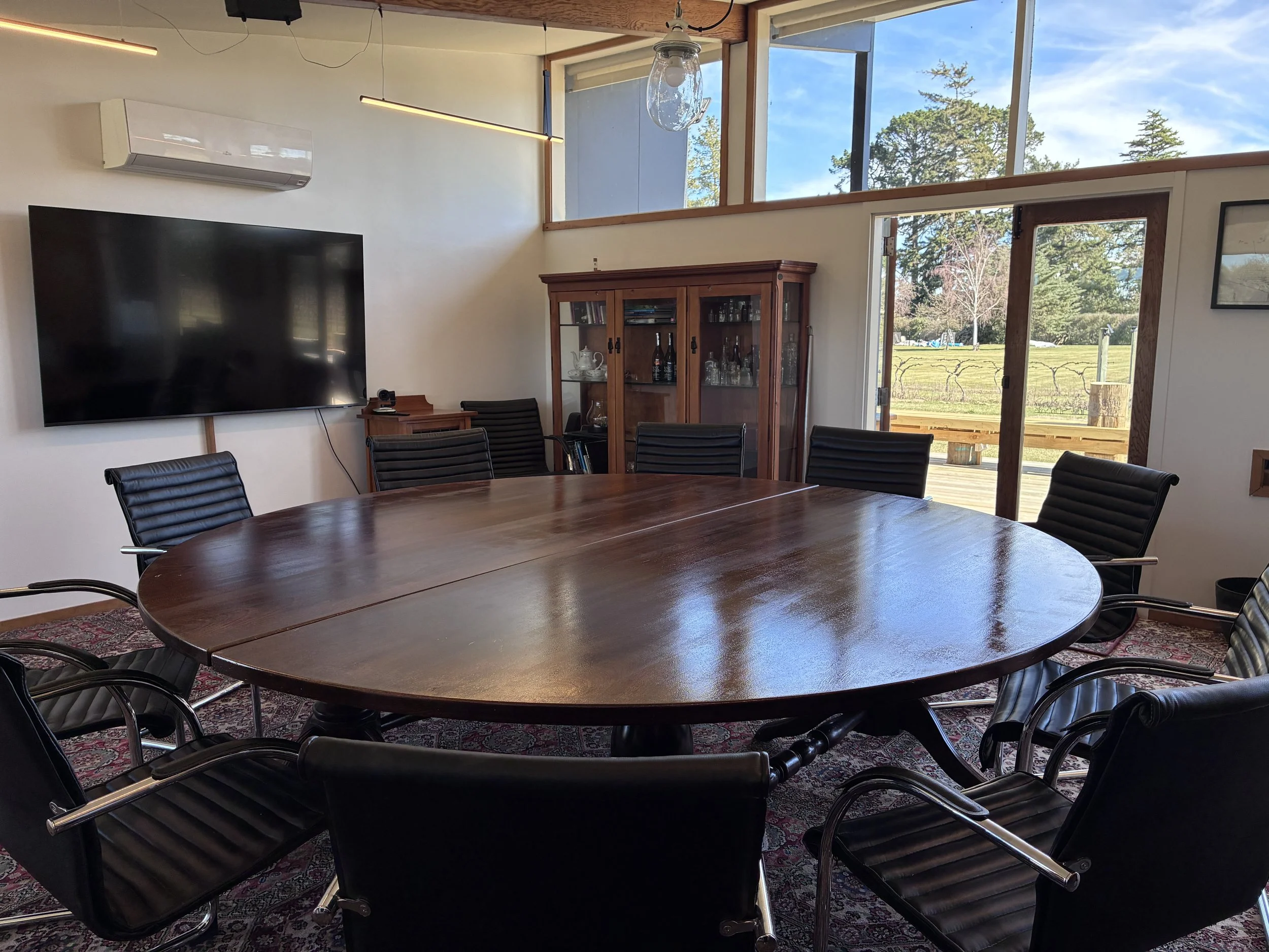 A conference room with a large oval wooden table surrounded by black chairs, a wooden cabinet with glass doors holding glassware and bottles, a wall-mounted flat-screen TV, windows showing trees outside, and french doors opening to an outdoor deck.