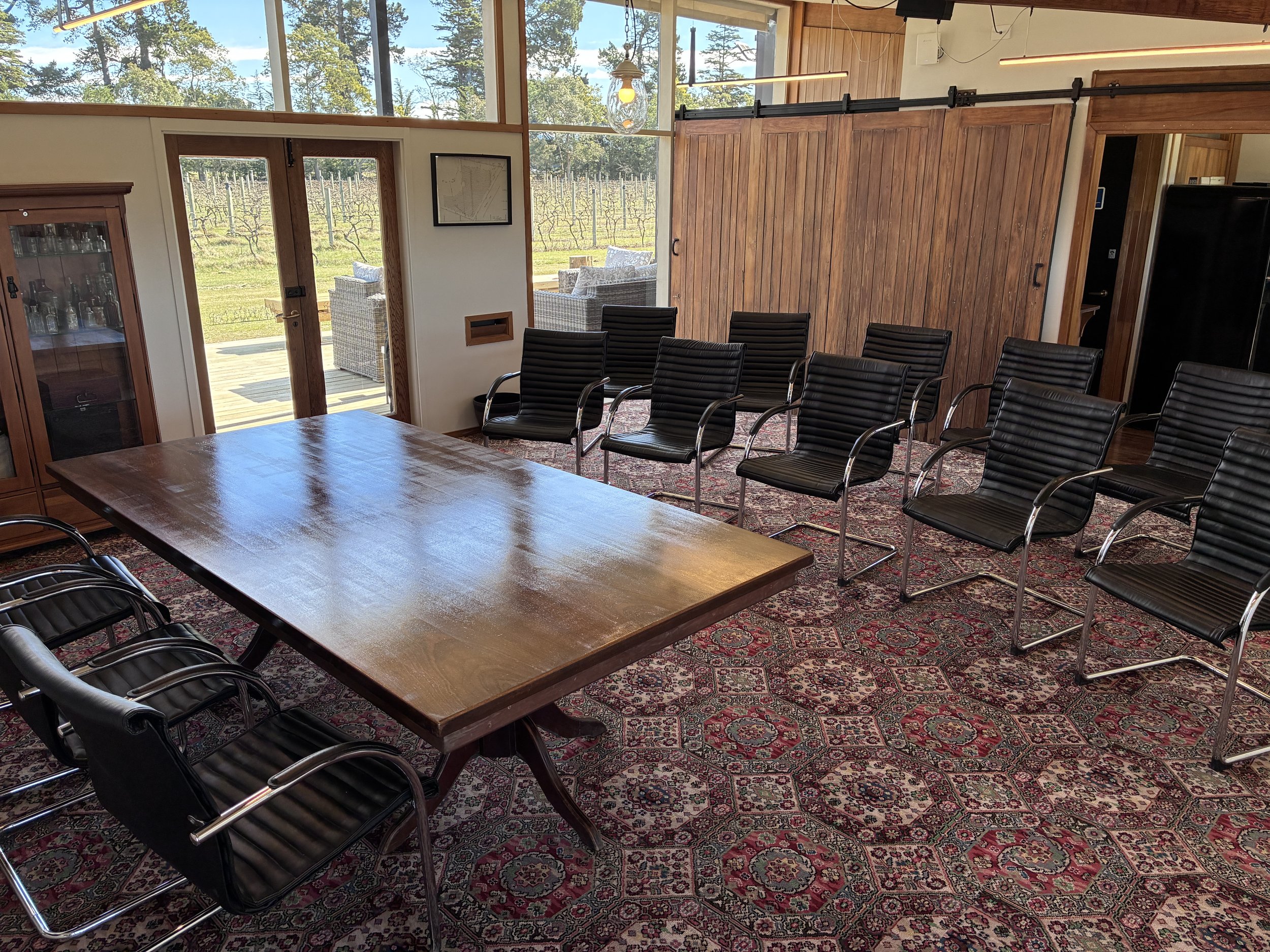 A conference room with black leather chairs arranged around a wooden table. Large windows show a view of a vineyard outside. The room has a patterned rug and wooden sliding barn doors.