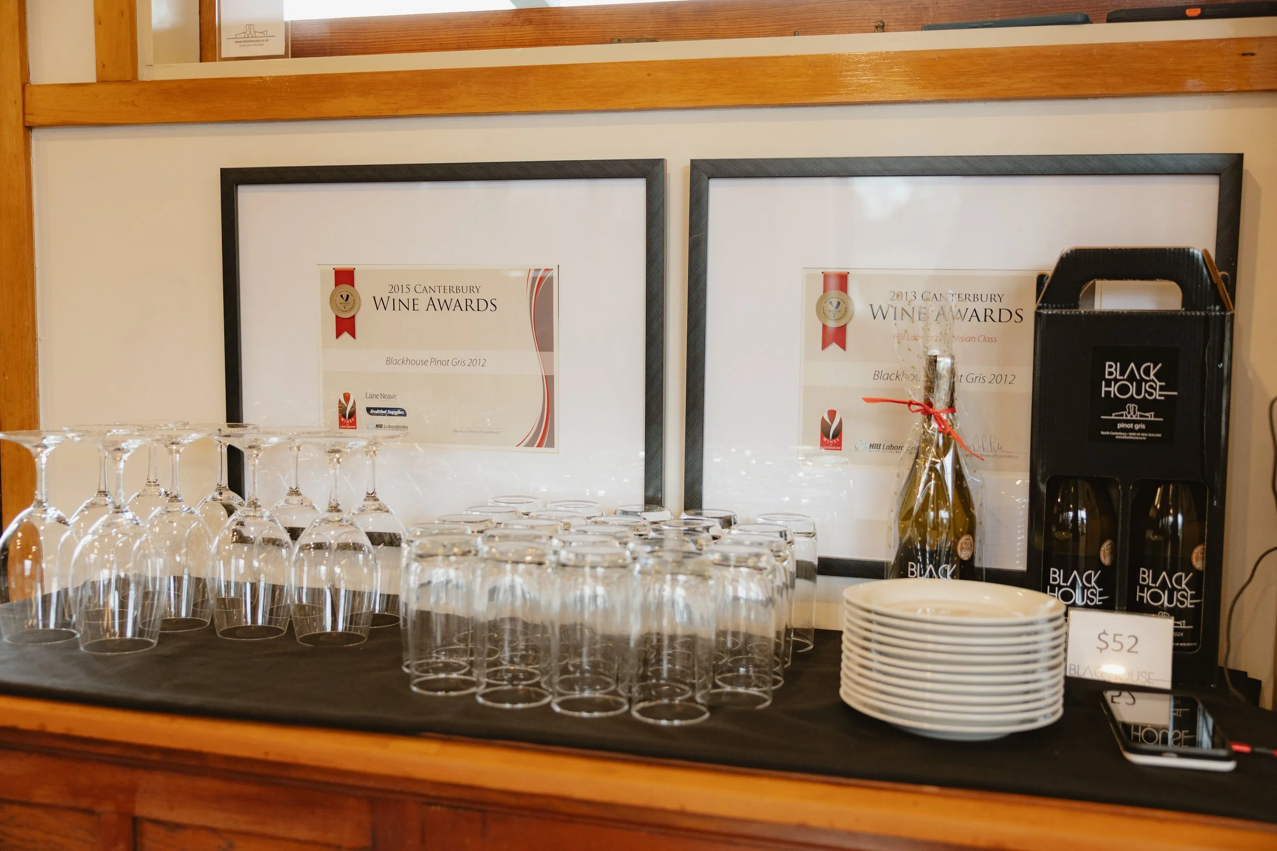 A display of empty wine glasses, framed certificates, and wine bottles at a wine awards event. The certificates are on a white background with red ribbons and gold seals, and the wine bottles are black with mostly white text. There are also glasses a
