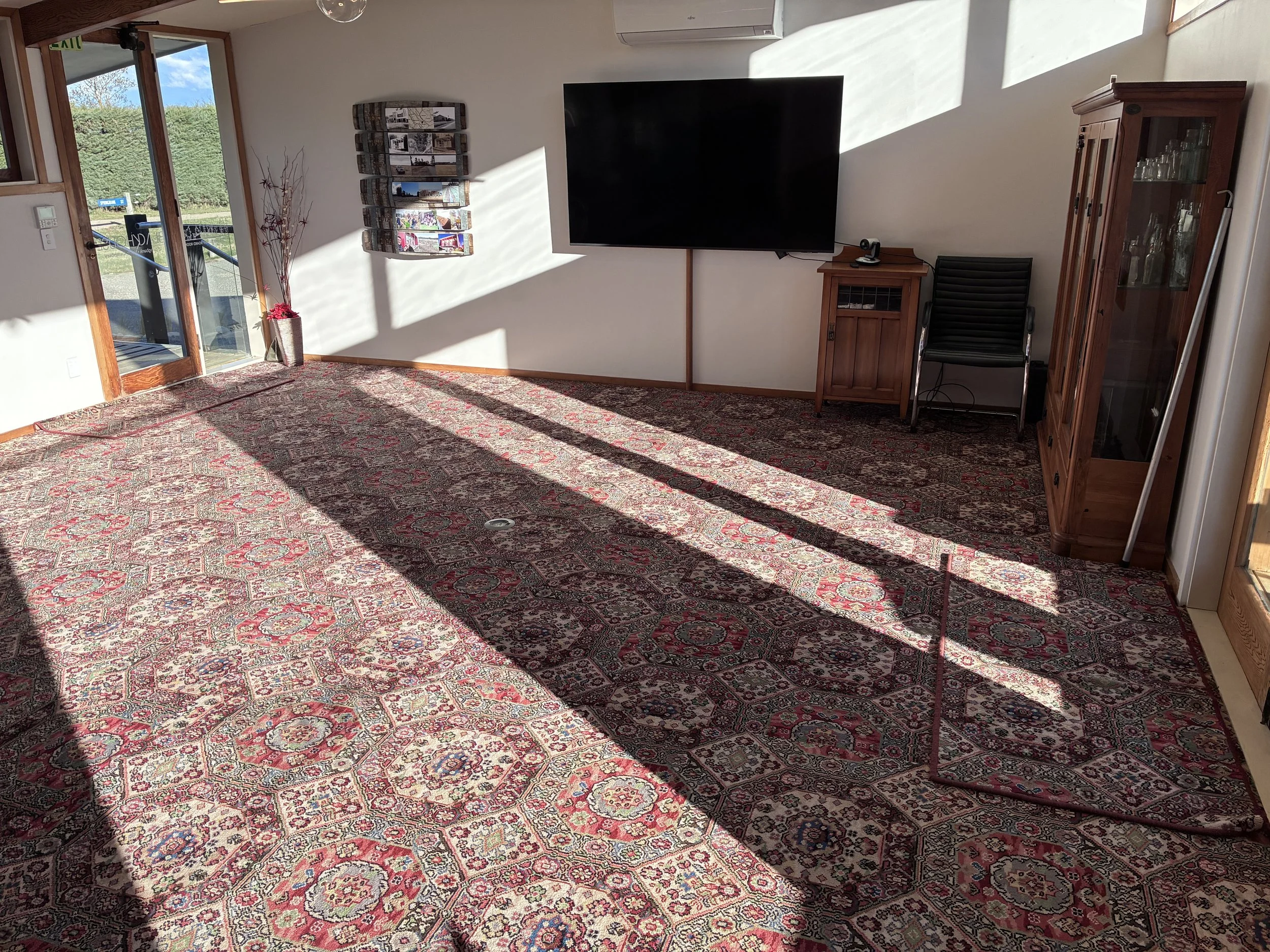 A living room with a large patterned rug, a wall-mounted TV, a wooden cabinet, a black chair, a glass-front display cabinet, a tall decorative vase with branches, a wall-mounted magazine holder, and an open door leading outside with a view of greener
