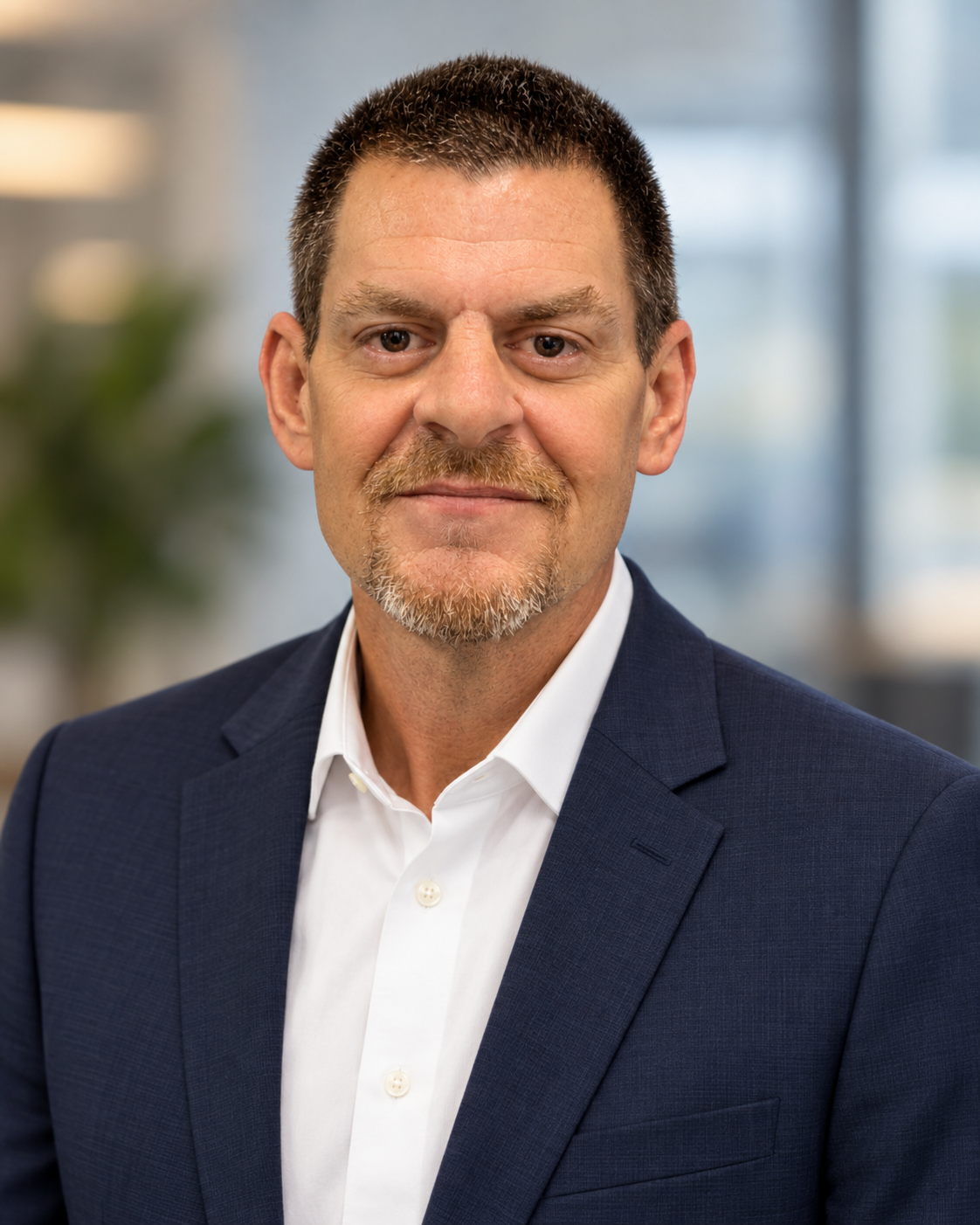 A middle-aged man with short dark hair, a mustache, and a goatee, wearing a dark blue suit and white dress shirt, smiling subtly in a professional headshot taken in an office setting.