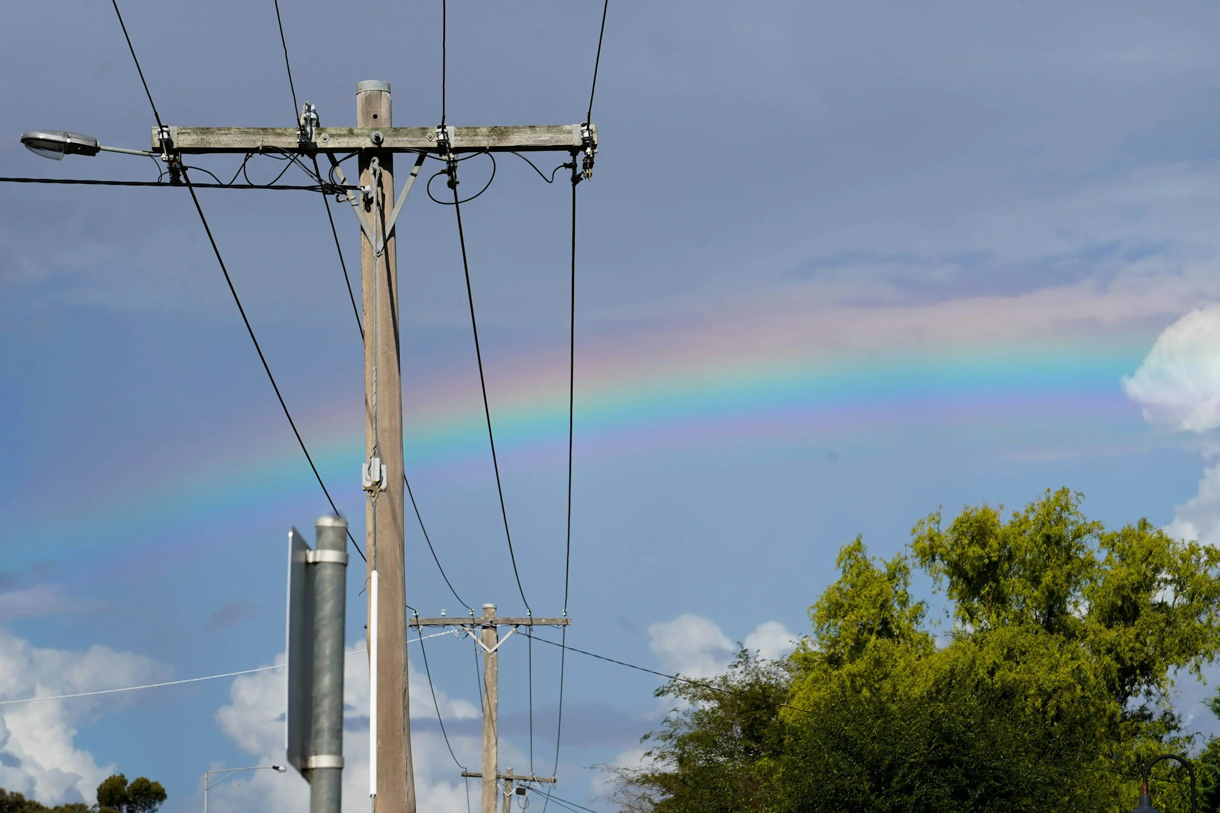 Electric power line poles with wires stretching across a partly cloudy sky with a faint rainbow and green trees in the background.