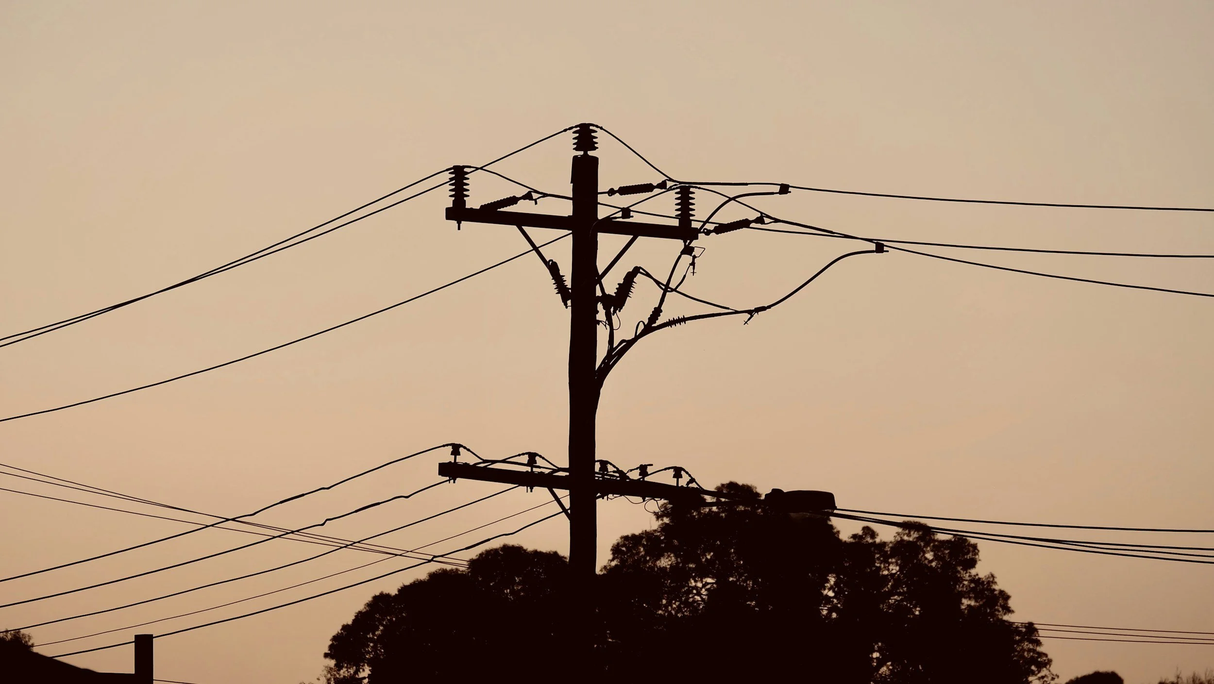 Silhouette of utility pole with multiple power lines against a dusky sky with trees at the bottom.