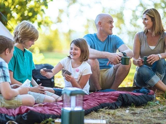 A family sitting outdoors on a blanket, playing cards and socializing in a park on a sunny day.