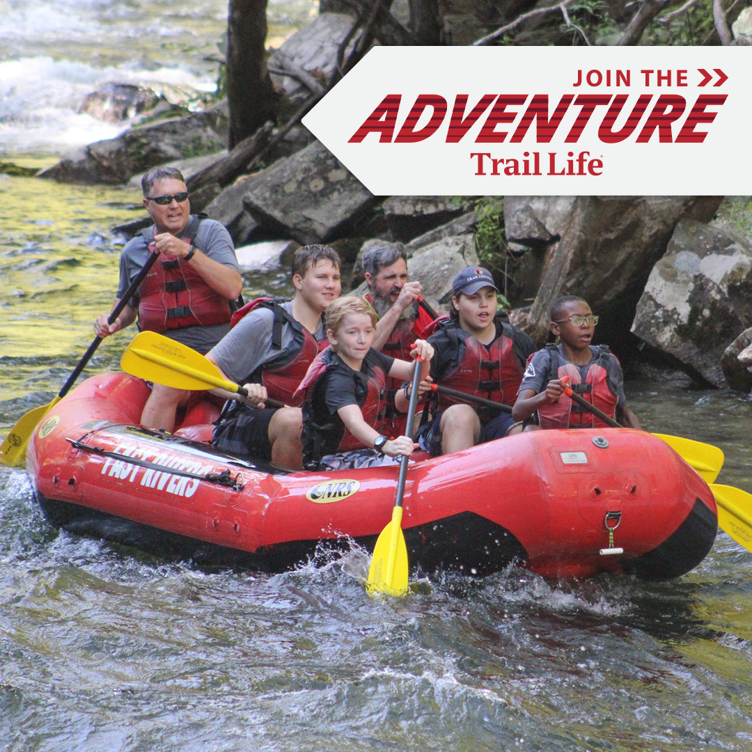 Group of six people white and African American in life jackets and outdoor gear, paddling a red inflatable raft adventure on a river surrounded by rocks and trees.