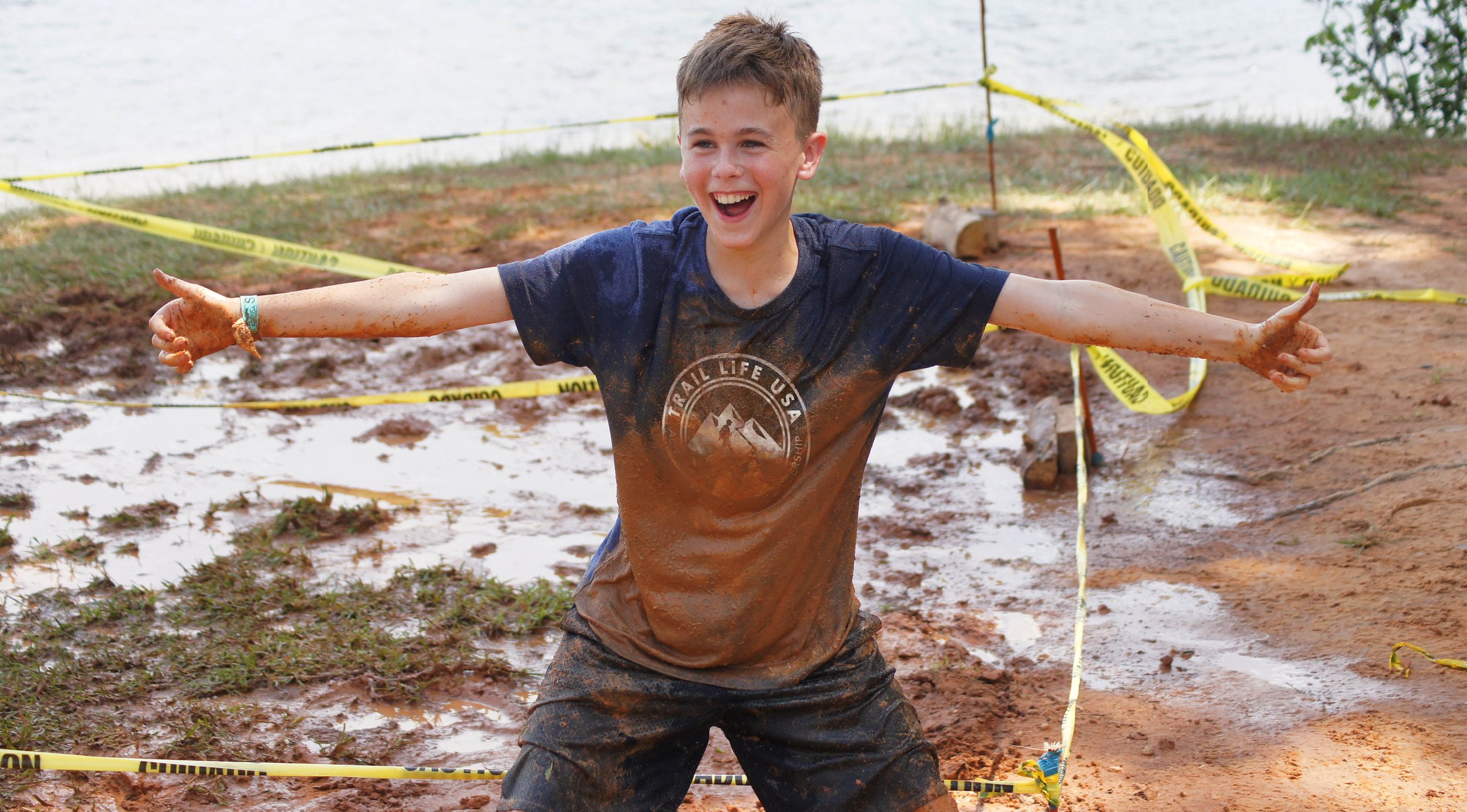 A young boy with dirty clothes and muddy arms, smiling with arms outstretched, standing in a muddy area marked with caution tape, near a body of water, celebrating after participating in a muddy outdoor activity or race.