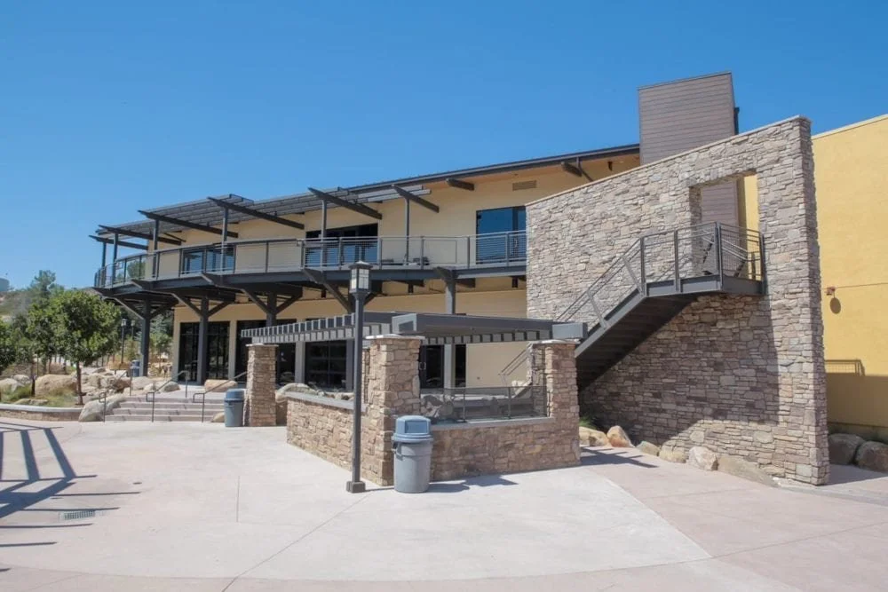 Modern building with stone walls and outdoor staircase, balcony, and seating area, under a clear blue sky.