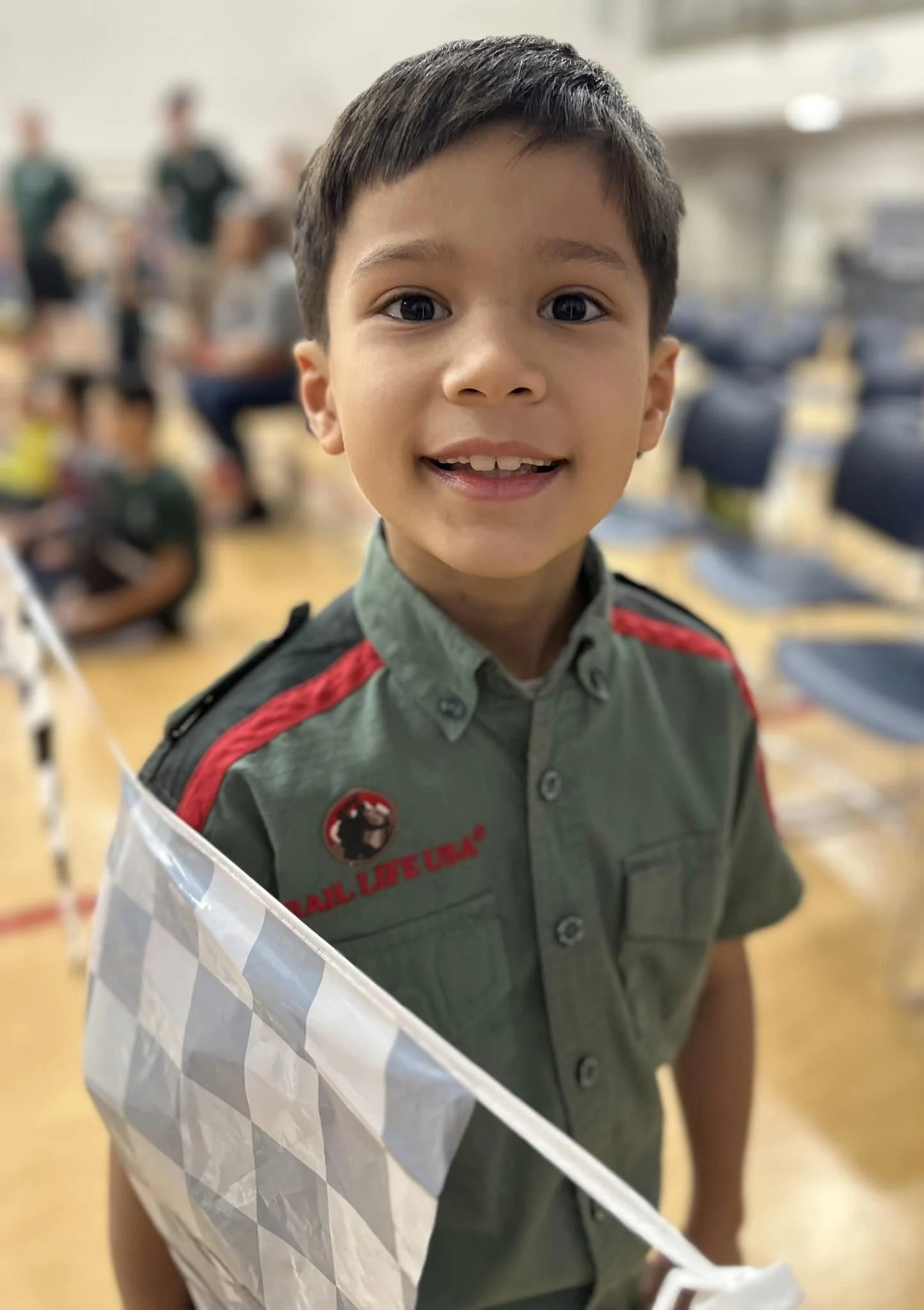 Young boy with short dark hair smiling at camera, standing in a room with other people in the background, wearing a green shirt with red and black patches, holding a checkered paper bag.