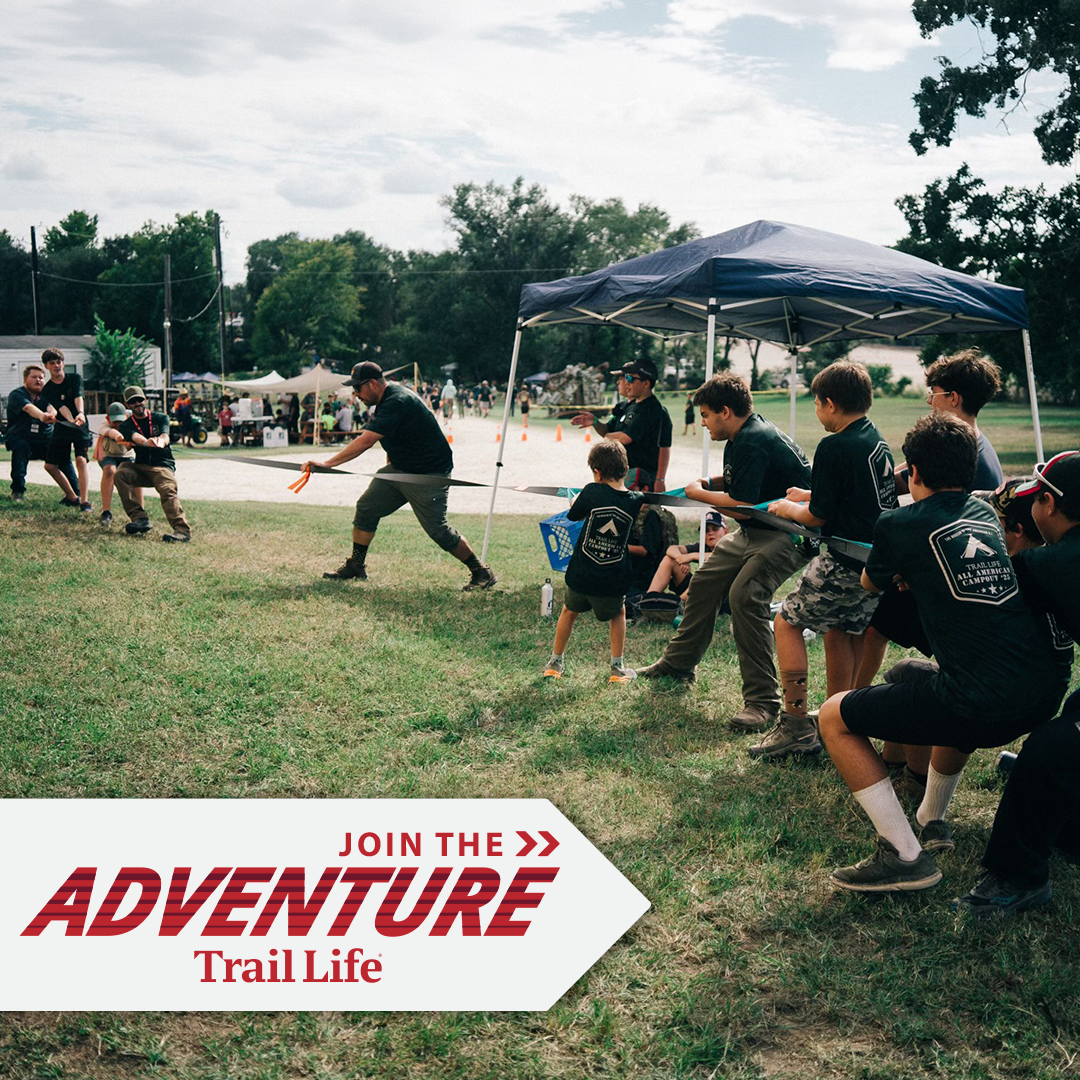Kids participating in a tug-of-war game at an outdoor event with tents and trees in the background, promoting trail life adventure.