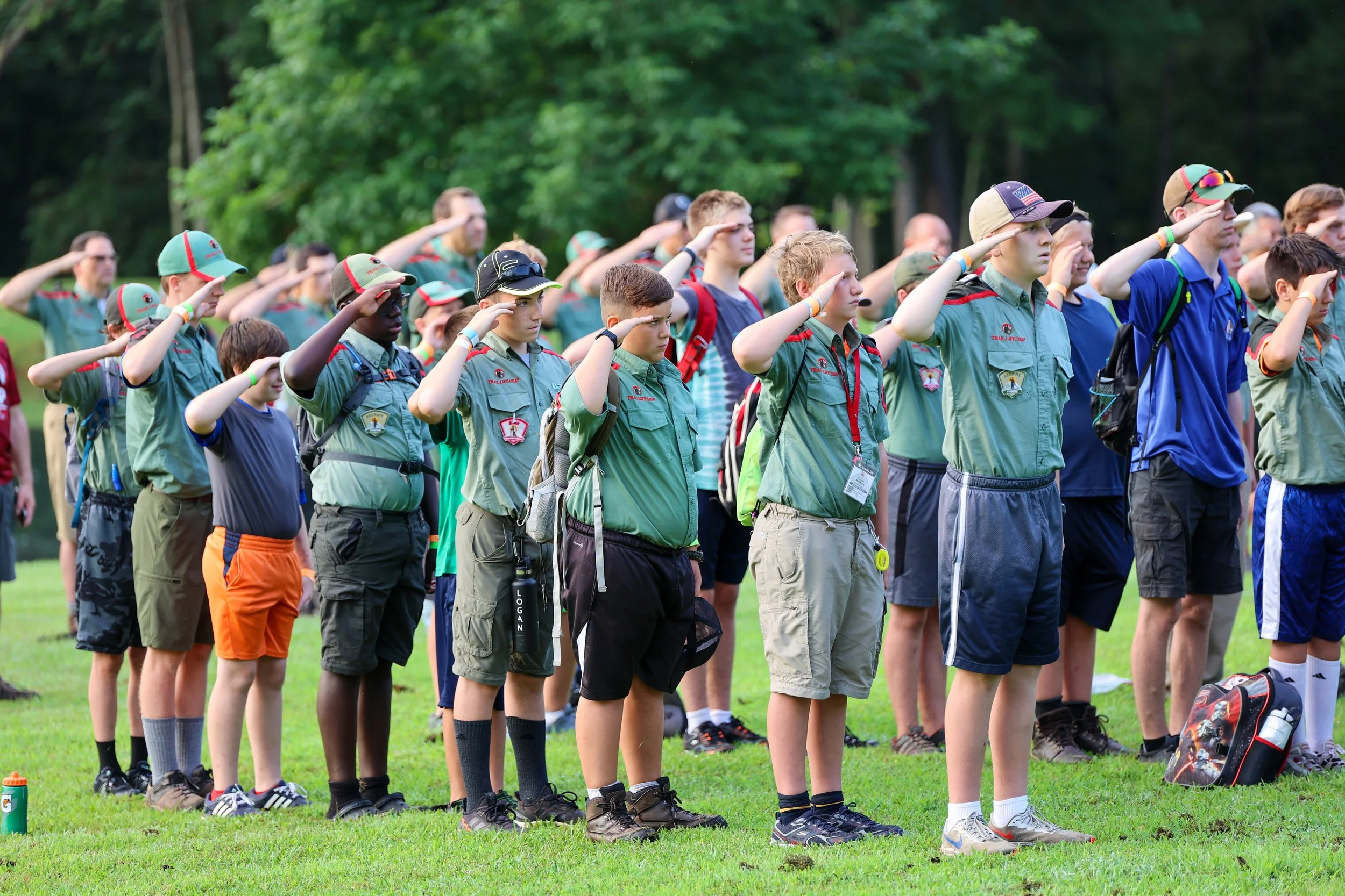 A group of boys and young men standing outdoors on grass, wearing Boy Scout uniforms and saluting during a ceremony or event.