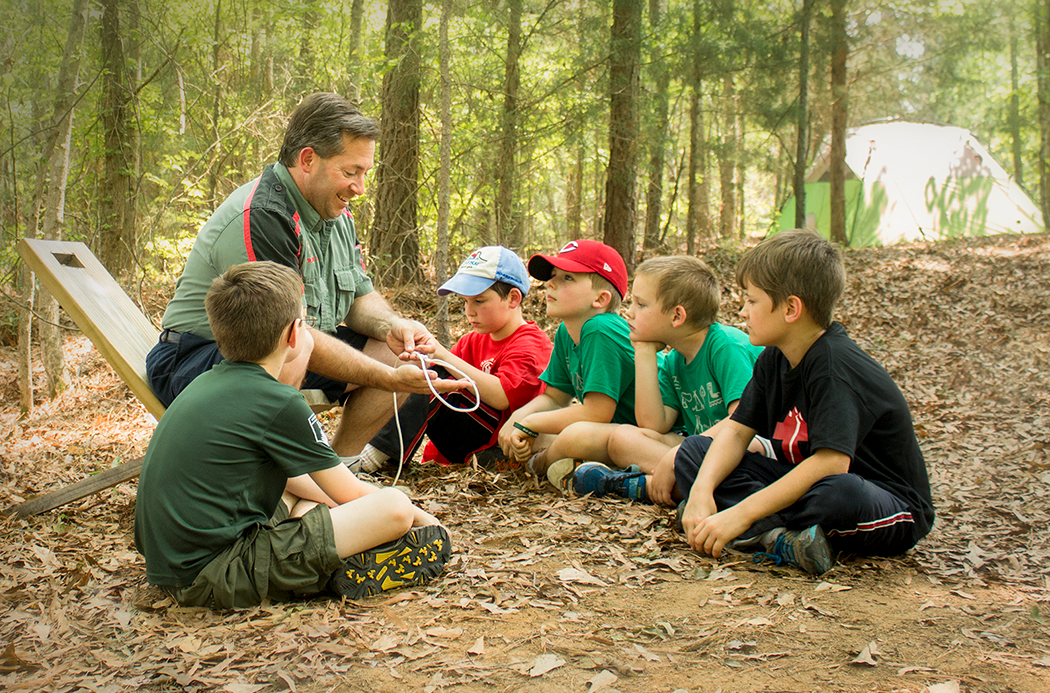 A man and five boys sitting in a forest, listening to the man demonstrate tying a knot.