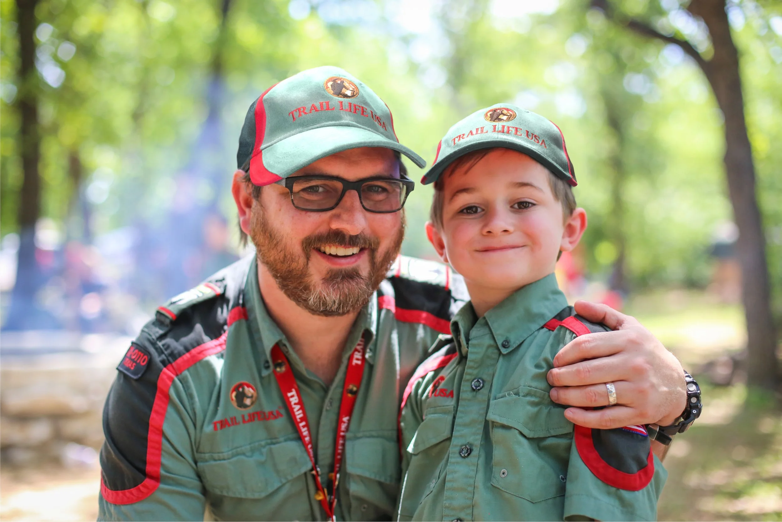 A man and a boy wearing matching green and red Scout uniforms and caps that read 'Trail Life USA' are smiling and hugging outdoors in a wooded area.