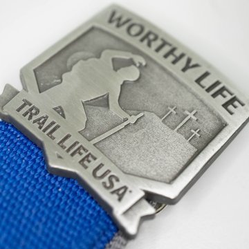 Close-up of a metal badge or pin with a silhouette of a hiker carrying a walking stick and a backpack, with the words "WORTHY LIFE" at the top and "TRAIL LIFE USA" at the bottom, attached to a blue fabric strap.
