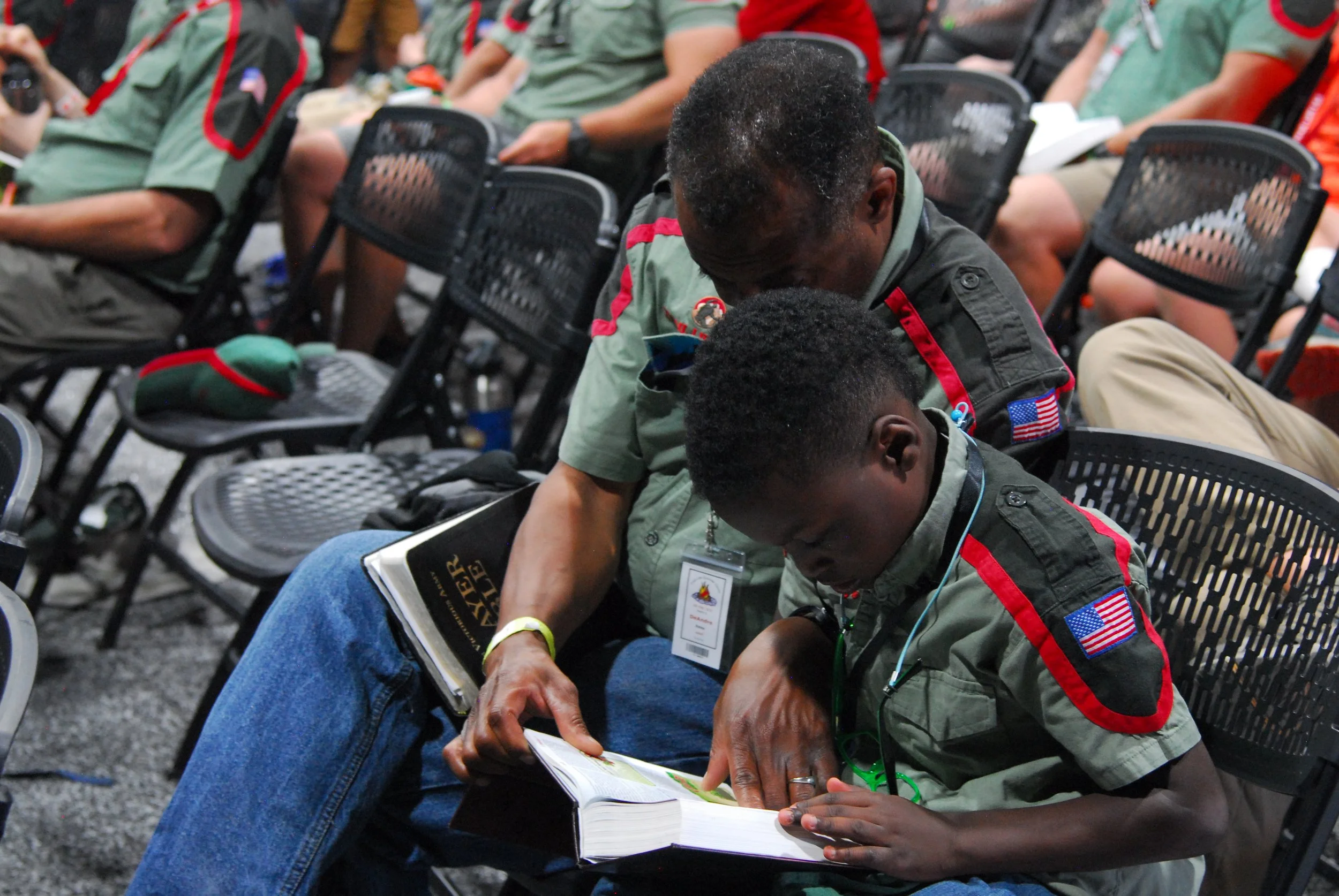 Military veterans in uniform sitting in chairs at an event, with one veteran reading a book while another looks on.