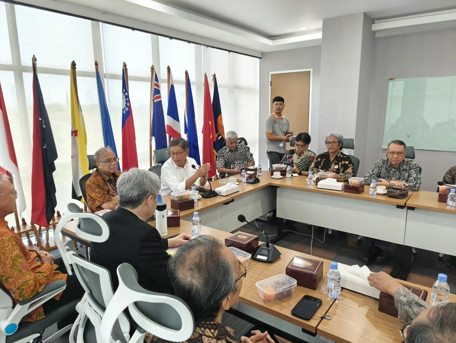 A diverse group of people sitting around a conference table in a meeting room, with flags of various countries in the background, some of whom are speaking and listening.