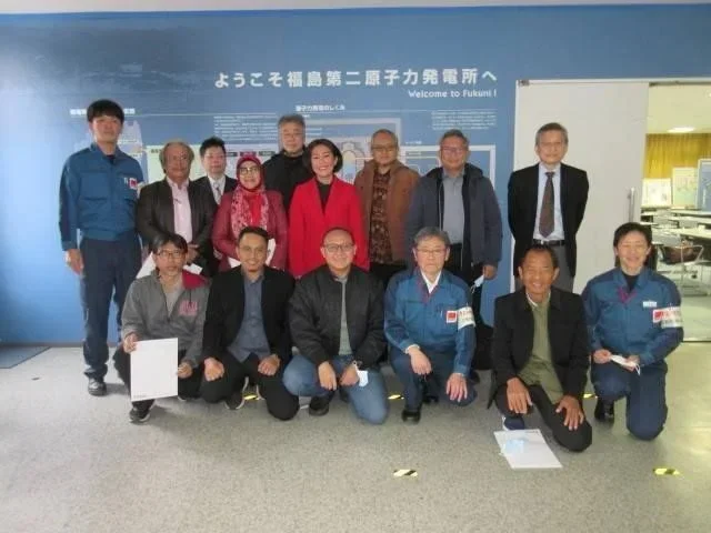 Group of men and women posing in front of a blue wall with Japanese writing and a welcome sign at a nuclear power plant.
