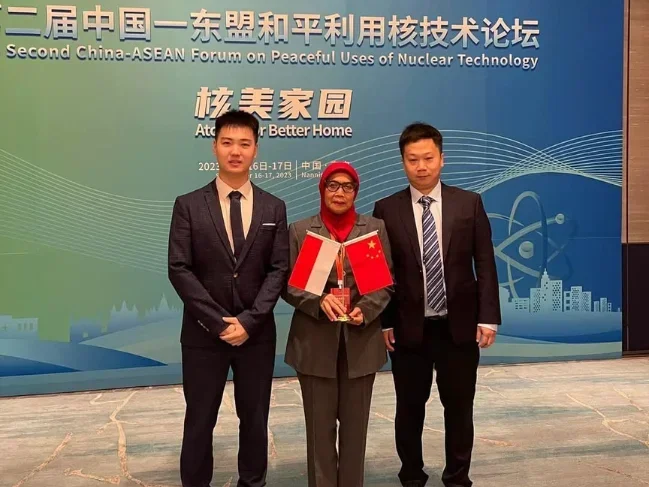 Three individuals standing in front of a blue backdrop at the Second China-ASEAN Forum on Peaceful Uses of Nuclear Technology, holding flags of China and possibly a related country, during a formal event.