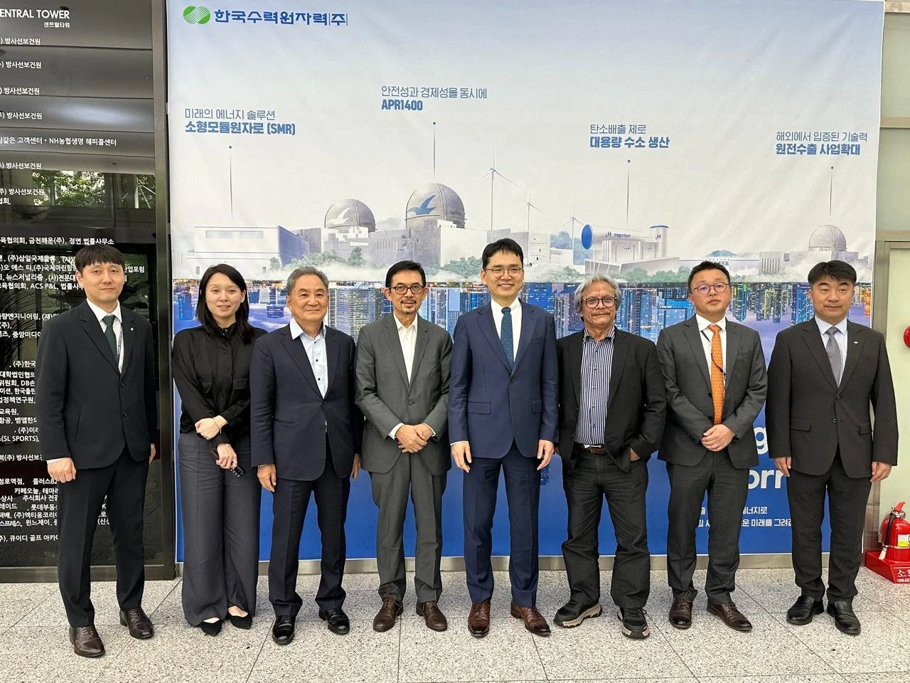 A group of nine business professionals standing in front of a large poster showing a nuclear power plant and text in Korean and English, posing indoors