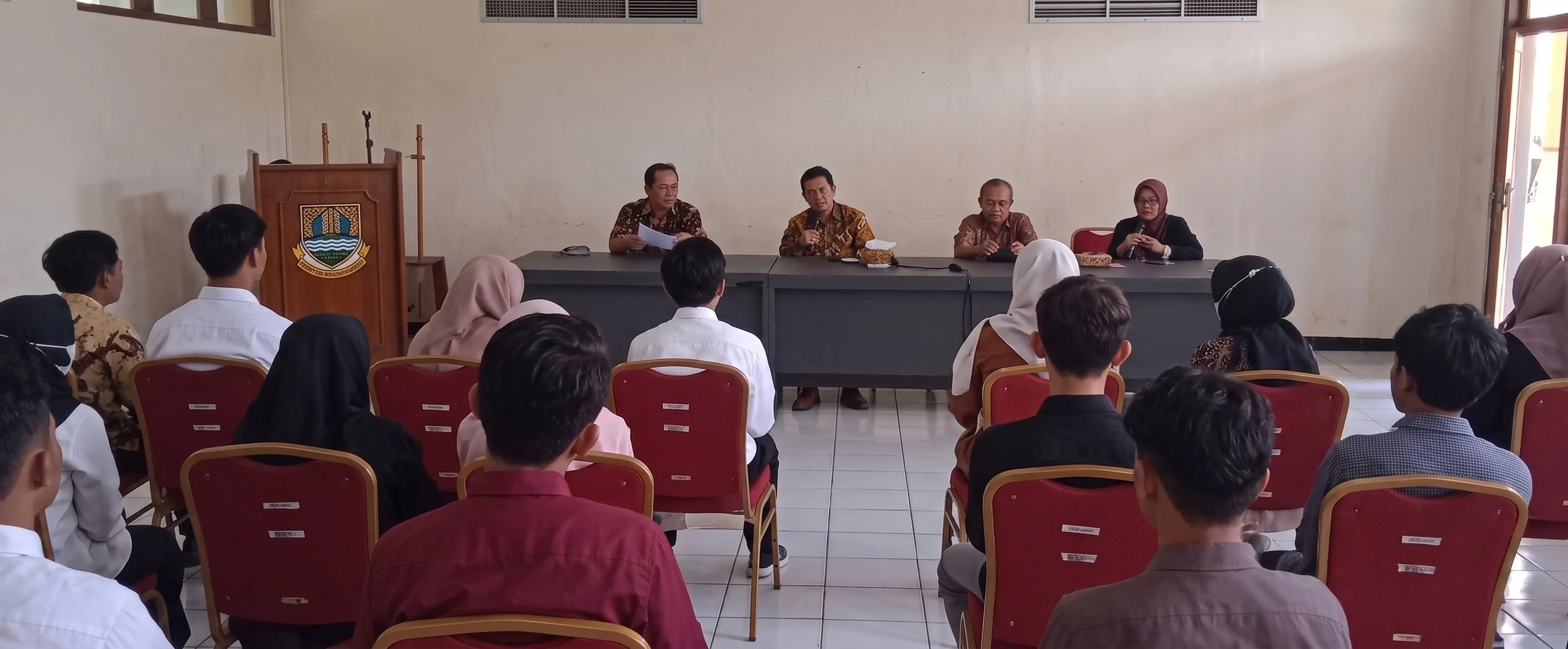 A classroom or meeting room with a panel of five people sitting at a long table at the front, facing an audience of students and adults, with some wearing batik shirts and others in white or formal attire. The room has plain walls, a door to the right, and an emblem or logo on a stand to the left.