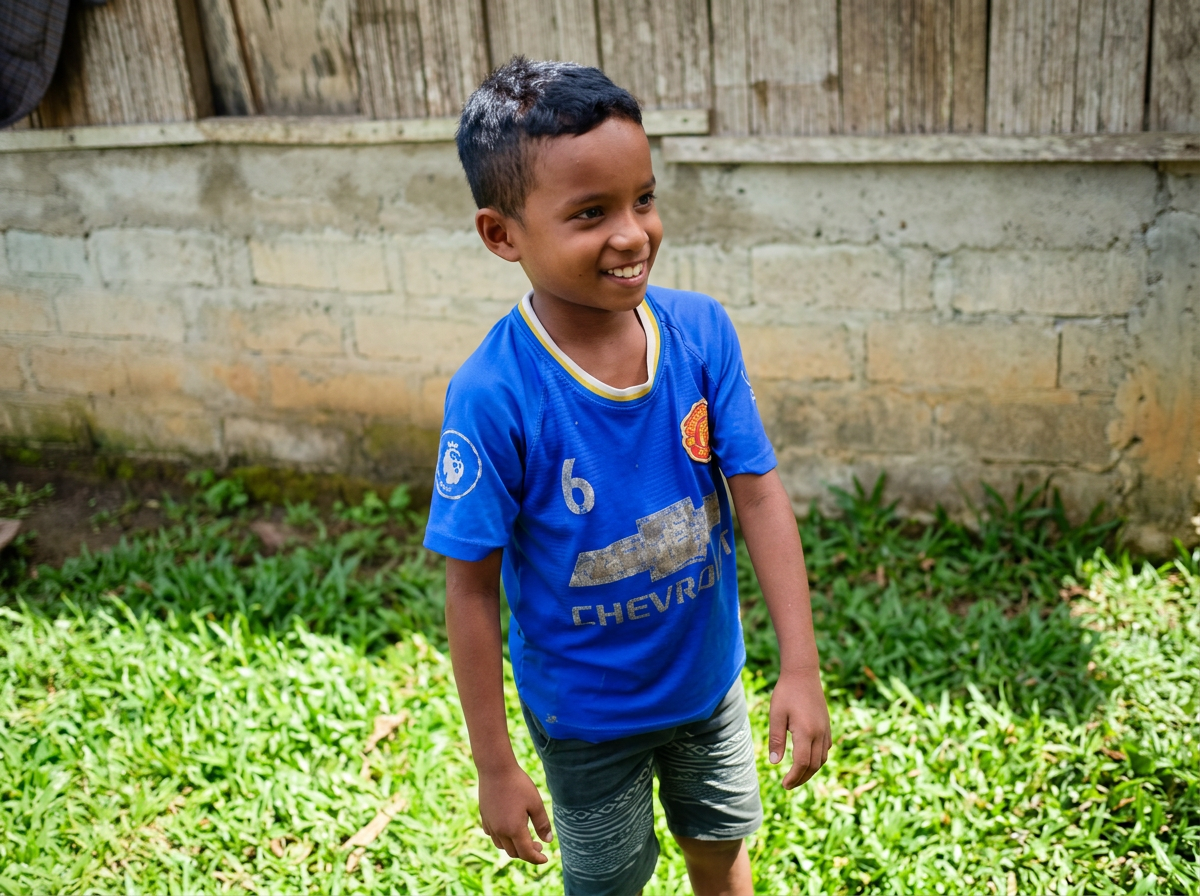 Smiling young boy with short black hair wearing a blue sports jersey and dark shorts, standing on grass in a backyard.