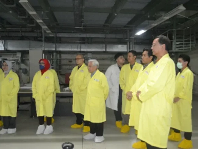 A group of eight people wearing yellow lab coats, face masks, and yellow shoe covers inside a lab or industrial facility, appearing to listen attentively.