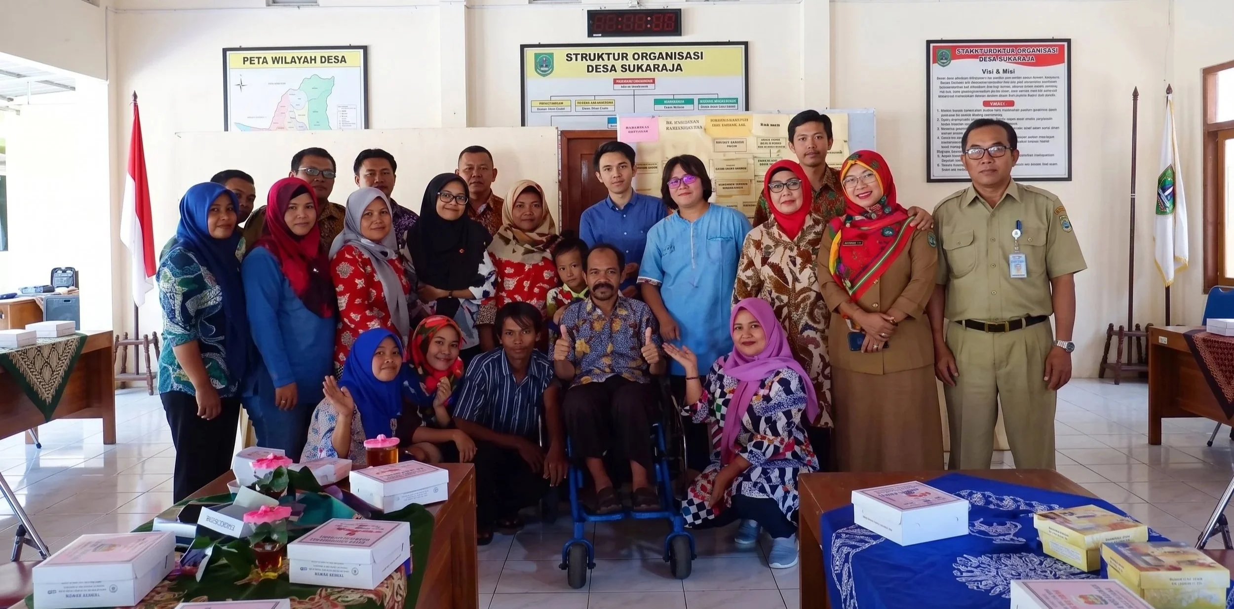 Group photo of adults and children inside a room with posters on the wall, Indonesian flags, and tables with boxes and flowers.